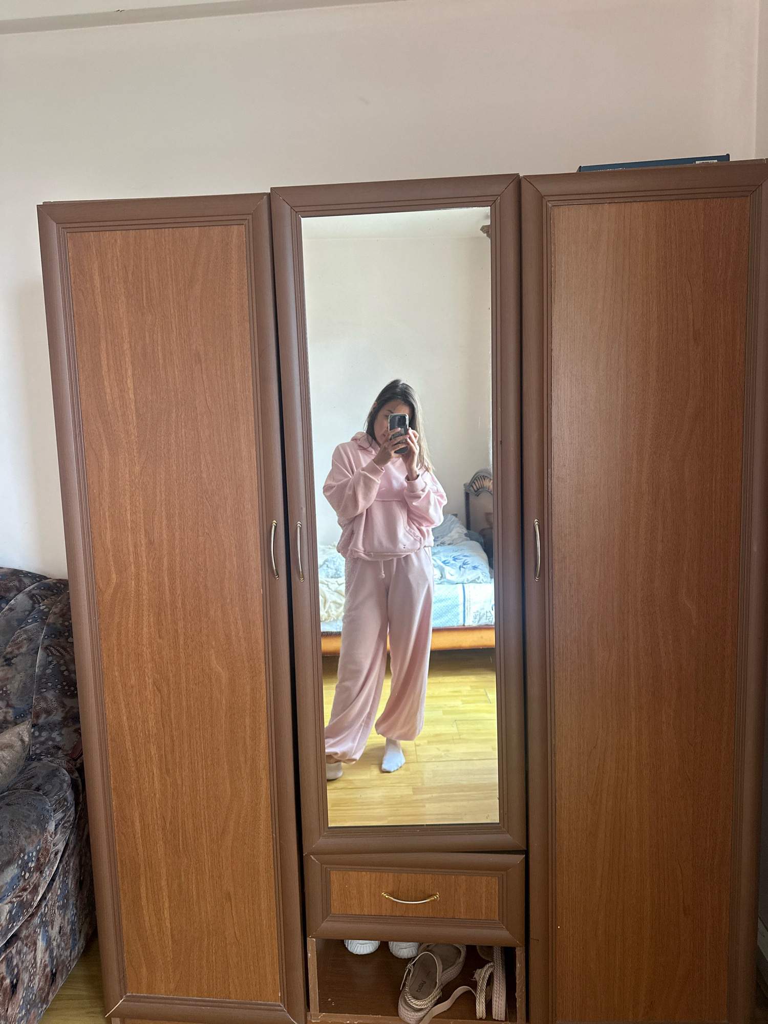 A young woman is taking a mirror selfie in a bedroom. She is wearing a pink hooded sweatshirt and matching pink pants. The woman is standing in front of a large, three-door wooden wardrobe with mirrored panels. The bedroom appears to be modest, with a bed visible in the reflection. The floor is wooden, and a portion of a patterned armchair is visible on the left side of the frame. The scene is illuminated by natural light. This image was taken in Gyumri, Armenia.