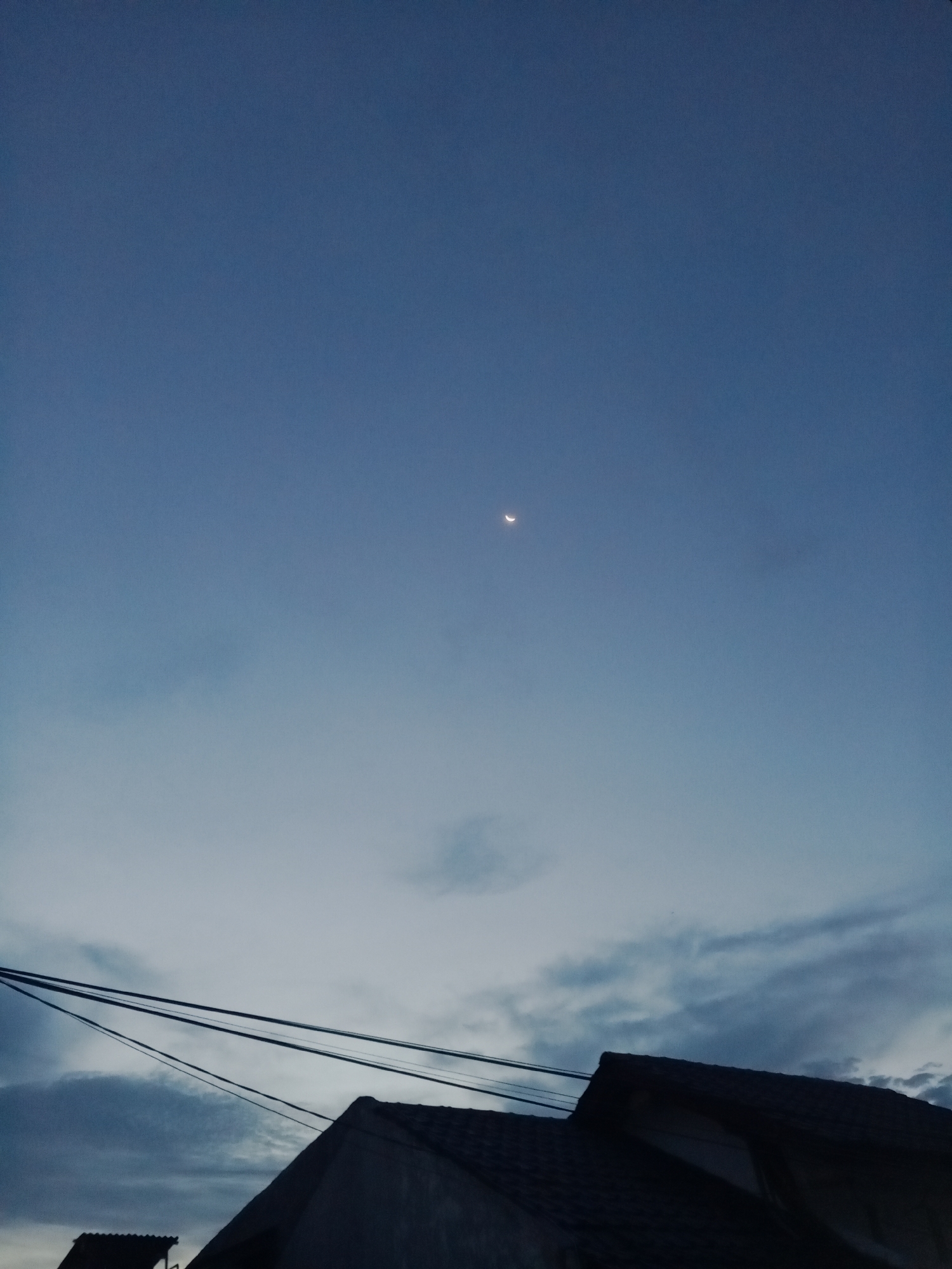 The image depicts a twilight sky over Lhokseumawe, Indonesia. A crescent moon is visible in the upper center of the frame. The sky transitions from a deep blue at the top to a lighter, cloudier gray closer to the horizon. In the lower portion of the image, rooftops of buildings are silhouetted, with power lines stretching across the foreground. The scene is still and absent of people.