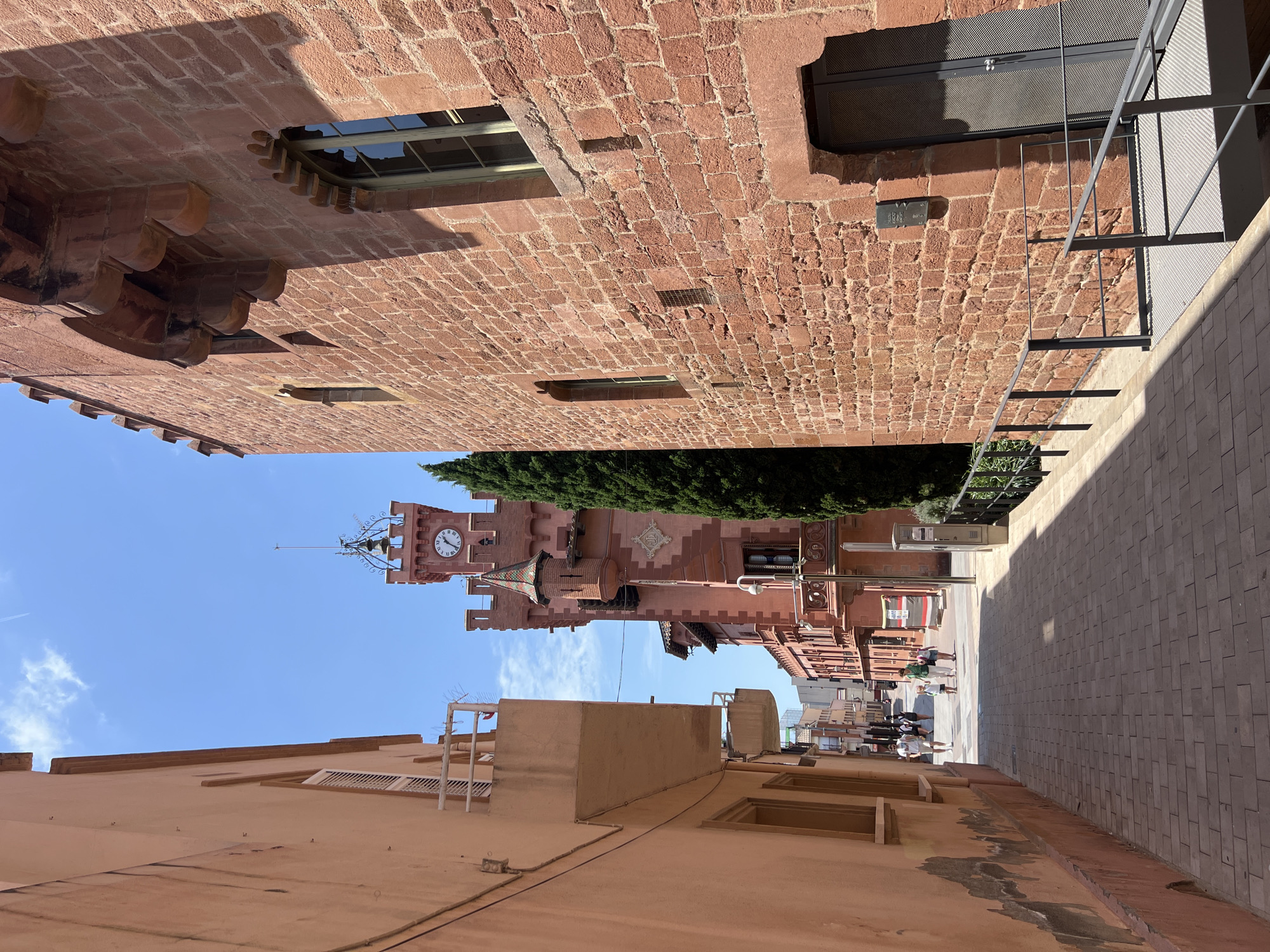 This is a vertical, eye-level, medium shot of a street in Viladecans, Spain, featuring two buildings. The building on the left is painted a pale orange and has a balcony. The building on the right is constructed of rough, red stone and has a decorative arched window and a large, metal-grated door. A narrow, paved walkway runs between the buildings. In the distance, a clock tower with a bell is visible. Several people are walking on the walkway. A tall, dark green tree stands next to the red stone building. The sky is blue with scattered clouds, and the sun is casting shadows.
