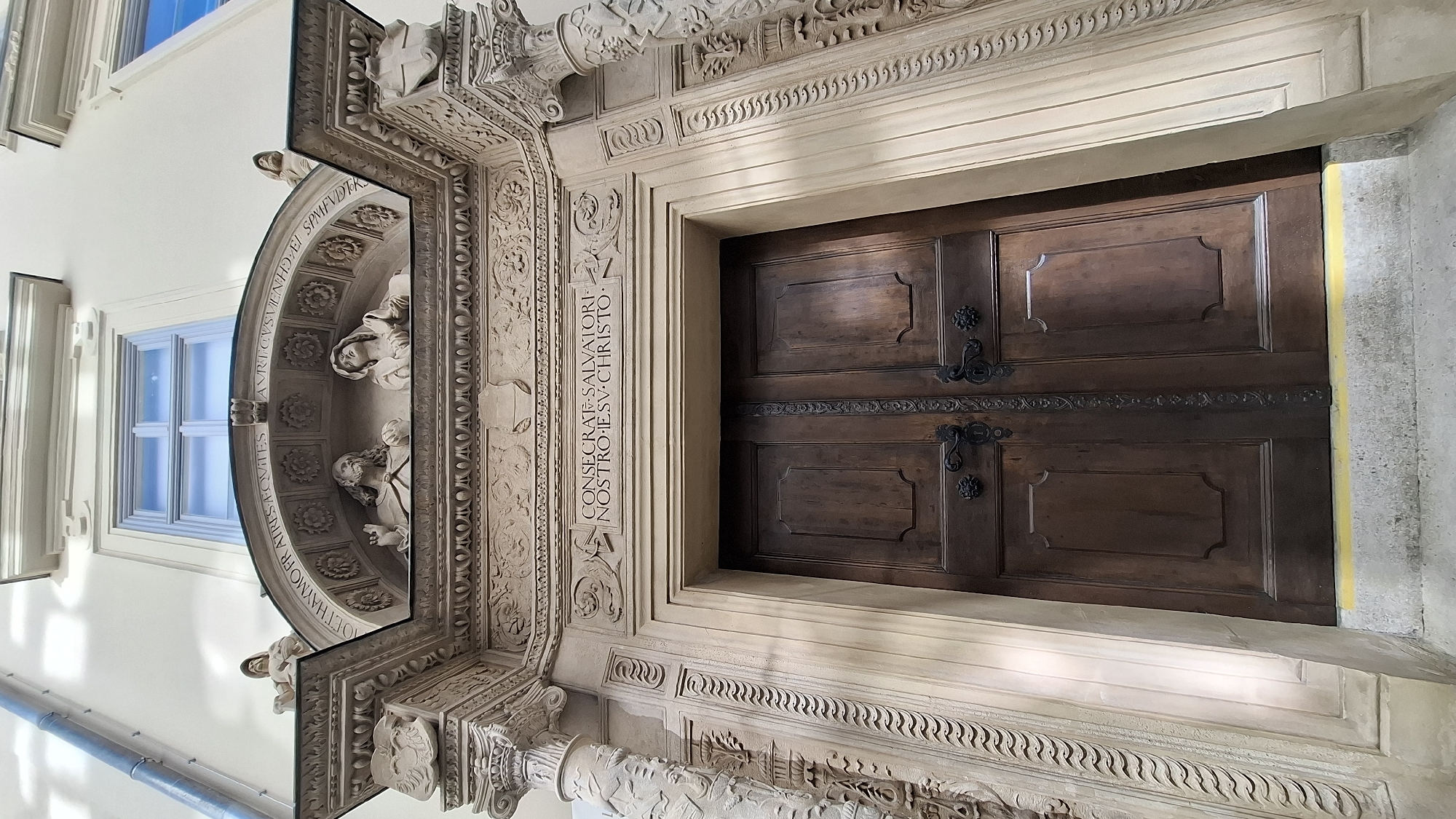 The image shows a detailed view of the main entrance to St. Stephen's Cathedral in Vienna, Austria. The entrance features a large, ornate wooden double door with dark metal hardware. Above the door, there is a stone inscription that reads "CONSECRATE SALVATORI NOSTRO IESV CHRISTO". The entrance is flanked by intricately carved stone columns and decorative stonework. Above the doorway, there is a sculpted pediment with figures and further decorative elements. A window with a blue sky visible through it is situated above the pediment. The architecture suggests a historical and religious site.