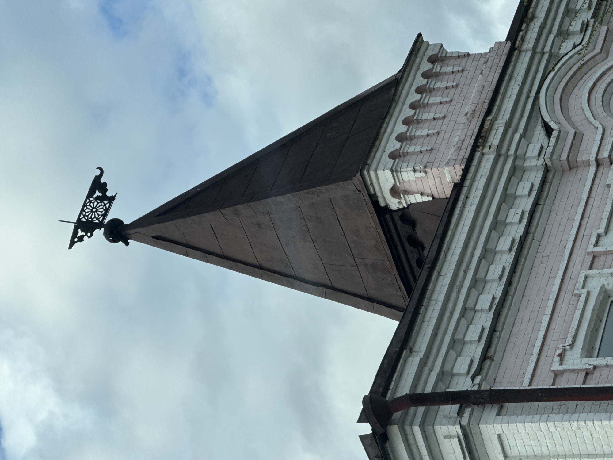 A low-angle shot depicts a building exterior in Syzran, Russia. The architectural style includes a steep, dark-colored conical roof topped with a decorative weather vane-like element. The adjoining building facade is pink and white, with detailed brickwork and architectural molding. The sky is overcast with gray and white clouds. A downspout runs along the side of the building.