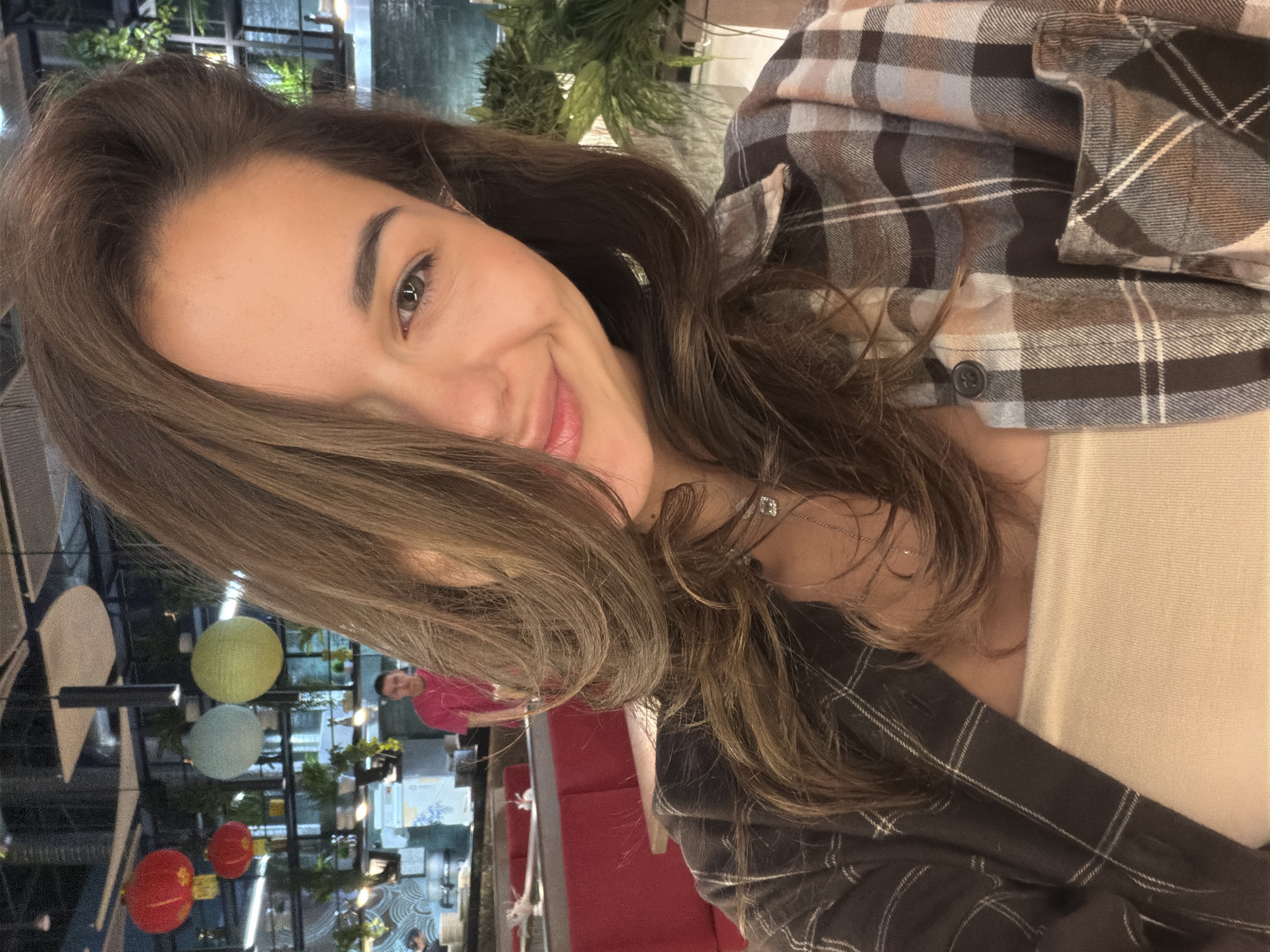 The image is a diagonal-oriented selfie of a young woman captured indoors, likely in a restaurant or cafe in Krasnoyarsk, Russia. She is looking directly at the camera with a bright, friendly smile, her long, wavy brown hair falling across her right cheek and shoulder. She has light brown eyes and well-defined eyebrows.
The woman is wearing a plaid button-down shirt featuring shades of brown, black, and light blue or pink, layered over a light, solid-colored (possibly beige or cream) top. A delicate silver chain necklace with a small pendant is visible around her neck.
The background, softly blurred, indicates an evening or night setting due to artificial lighting. It features modern interior elements including dark structural beams, lush green foliage, and various decorative spherical lanterns or orbs in yellow, light blue, and red. Red upholstered booths or seating can be seen, and a person in a bright pink top is visible further back in the background, suggesting a social environment. The overall atmosphere appears relaxed and inviting. There is no visible text in the image.