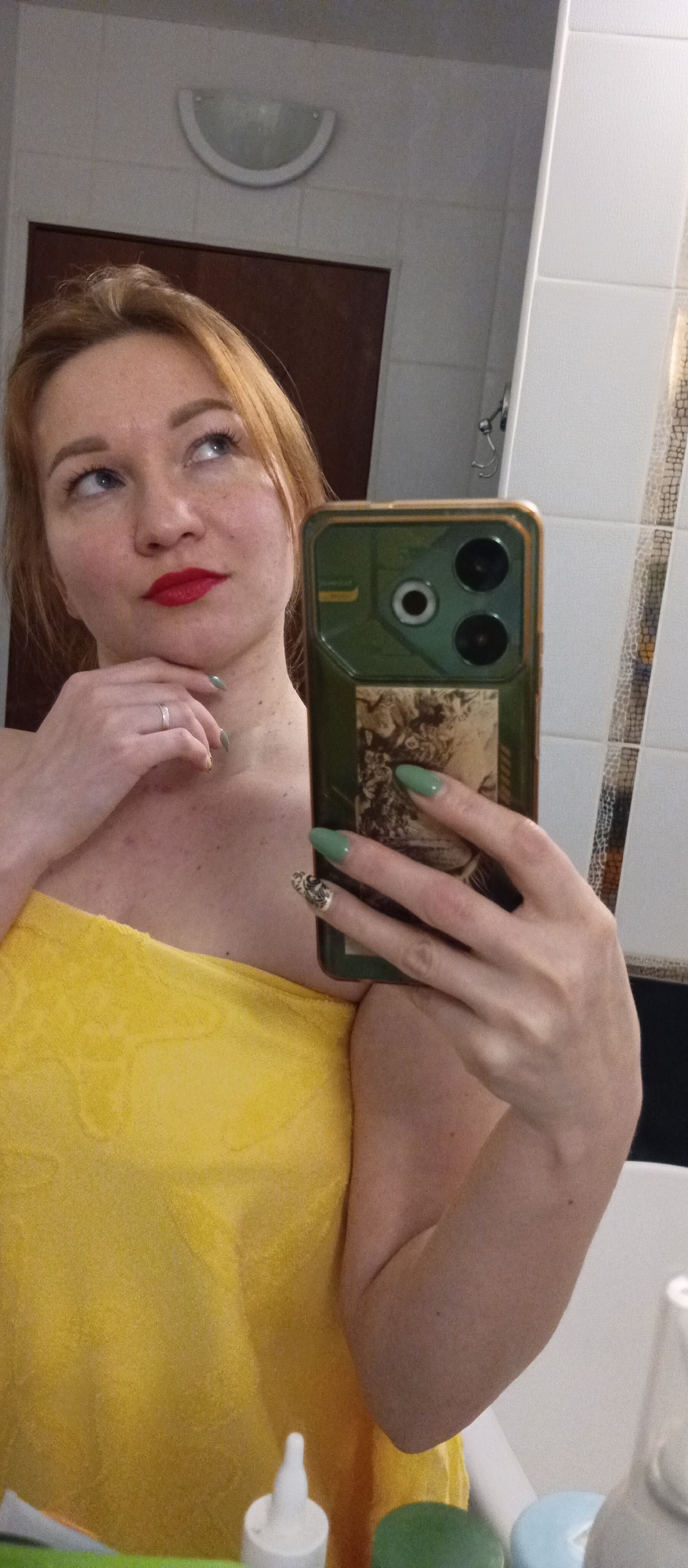 A woman wearing a yellow towel is taking a selfie in a bathroom. The bathroom has white tile walls. The woman is looking upwards with her right hand resting on her chin. She is wearing red lipstick and has green nail polish. In her left hand, she holds a smartphone with a green case and three camera lenses. Below the phone, a section of a bathroom counter is visible with several bottles. The location is in an unknown city.