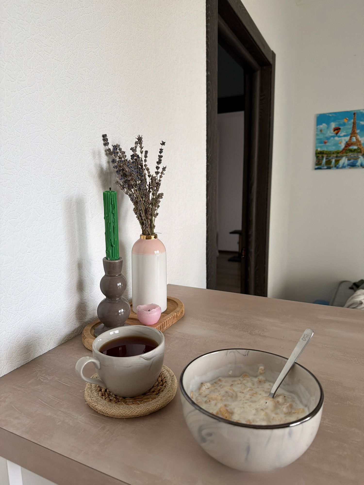 An indoor scene, likely a dining area in Kyiv, Ukraine, shows a table with breakfast items and decorative objects. On the left, a textured white wall forms the background. A dark brown wooden doorframe is visible on the right, with a glimpse of an interior space. A small painting of the Eiffel Tower with hot air balloons hangs on the wall.
On a beige surface, a light grey mug filled with dark liquid sits on a woven coaster. Next to it, a bowl of cereal with milk is placed, with a silver spoon resting inside. To the left of the mug, on a wooden tray, are decorative items: a dark grey ceramic candle holder with a tall, dripping green candle, a pink candle in a small dish, and a white vase with a pink top containing dried lavender. The lighting appears to be natural, coming from the right. No people are present in the scene.