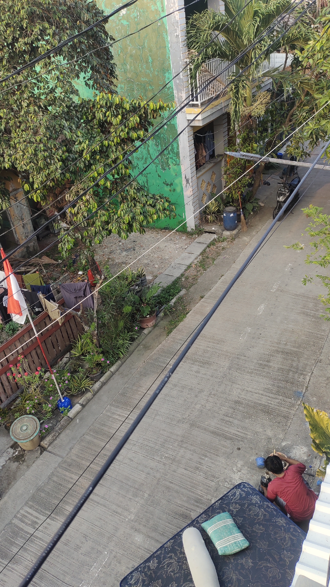 In Jalenjaya, Indonesia, a residential scene unfolds along a paved street bordered by a narrow dirt path and a low wooden fence. A person, wearing a red t-shirt and dark pants, is seated on a mattress on the ground, facing away from the camera and engaged in tending to a small plant. Adjacent to them, a blue bucket and a broom rest on the ground. Further down the street, a motorcycle is parked. The environment is characterized by a two-story building with a green facade and a balcony, surrounded by lush vegetation. Laundry hangs on a clothesline in front of the building. Overhead, a network of electrical wires crisscrosses the scene.
