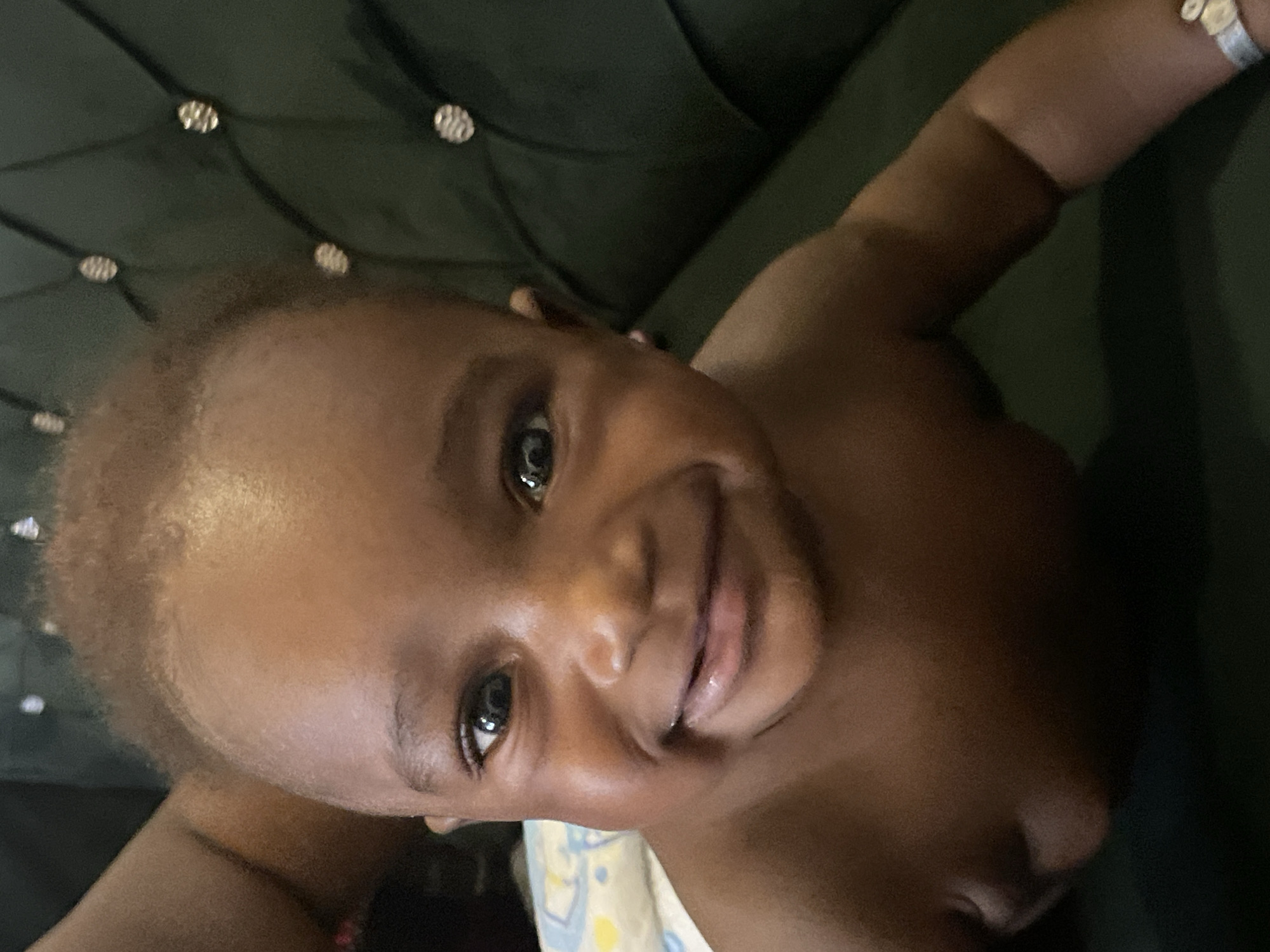 A close-up, slightly angled shot of a young child, likely an infant or toddler, with dark skin and curly light-brown hair. The child is lying on their back against a dark green, button-tufted surface, possibly a couch or headboard. The child's face is the focal point, displaying a small smile. The child has dark eyes and slightly parted lips. A white hospital identification band is visible on the child's right wrist. The child appears to be wearing a diaper, partially visible at the bottom edge of the frame.
This image cannot be confirmed as being taken in Barakin Akawo, Nigeria, as the location is not discernible from the visual information provided.
