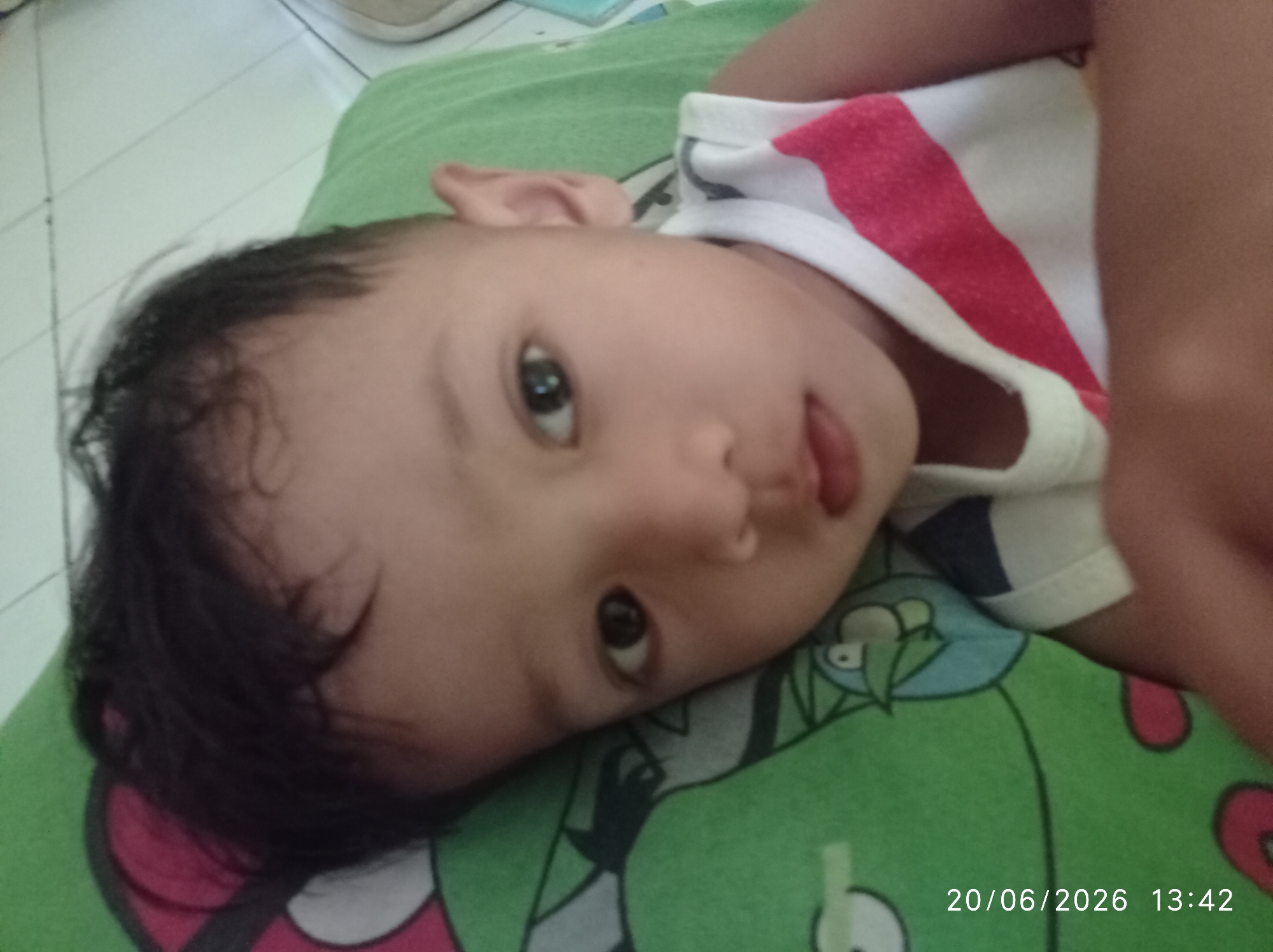 A close-up, slightly angled shot shows a baby lying on a green blanket with a cartoon character print. The baby's head is turned to the side, with dark hair and large, expressive eyes. The baby is wearing a white t-shirt with red and blue stripes. A portion of the baby's arm is visible on the right side of the frame. The overall setting appears to be indoors, with a tiled floor visible in the background. The timestamp "20/06/2026 13:42" is visible in the bottom right corner of the image, suggesting the photo was taken on June 20, 2026, at 1:42 PM. The lighting suggests it is daytime. The baby's facial expression is curious and engaging. The location context is Sukaraja, Indonesia.
