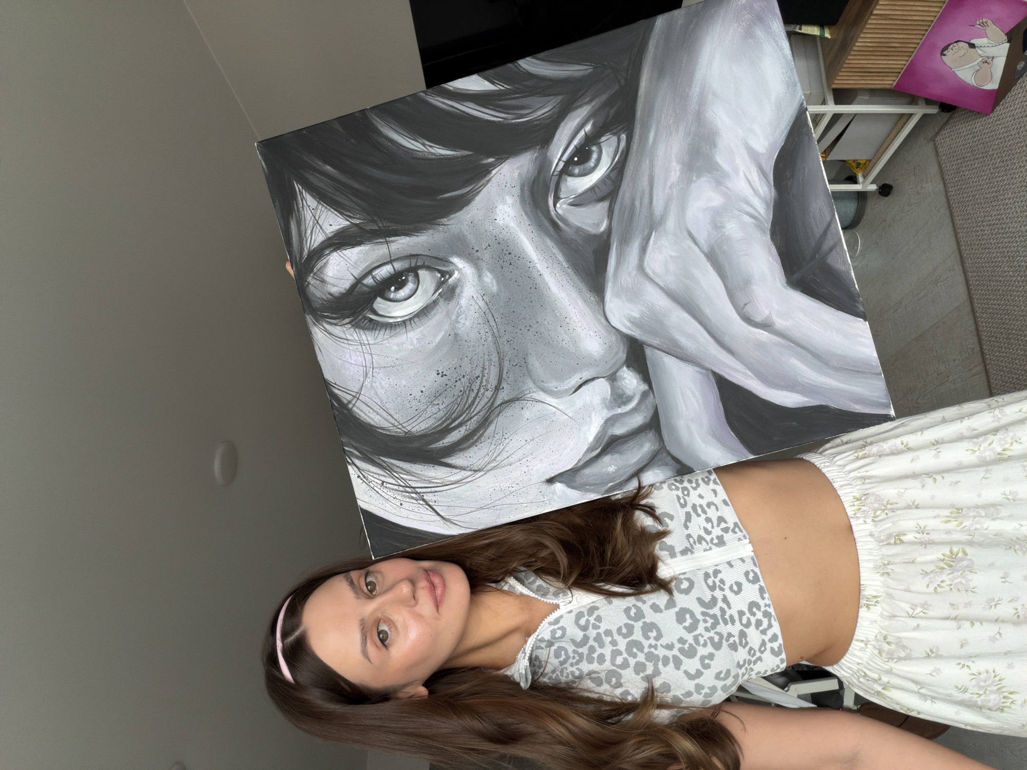 A young woman with long brown hair, wearing a light-colored, patterned crop top and a floral skirt, holds a large black and white painting. The painting is a close-up portrait of a person's face, focusing on their eyes and hands. The woman is standing indoors, in what appears to be a living space, with a ceiling light visible above. The background shows a wall and a shelf unit. There is no specific indication of the location being Krasnoyarsk, Russia.