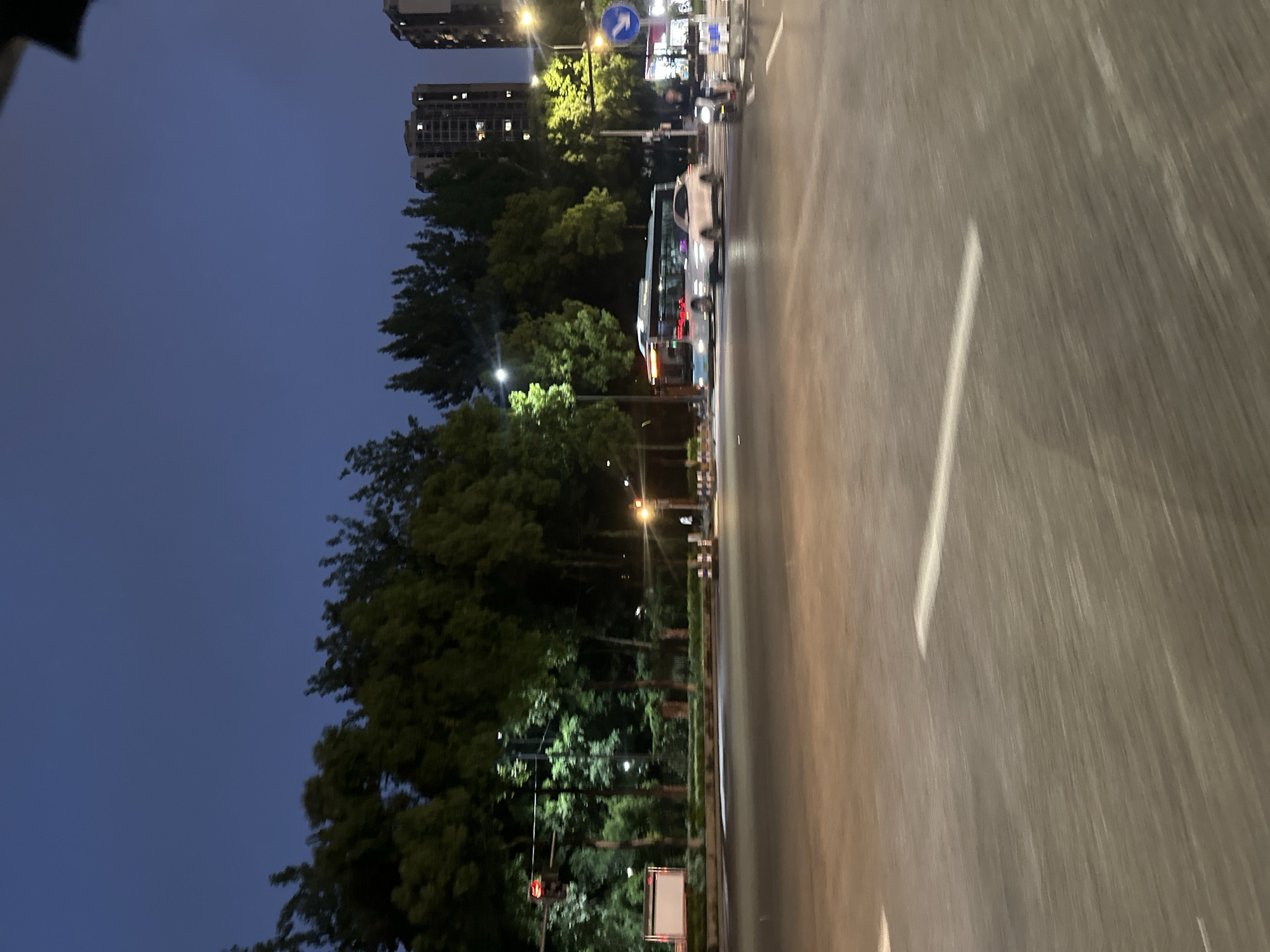 This is a medium shot of a street scene at dusk in Chongqing High-tech Zone, China. The sky is a deep blue, transitioning to lighter hues near the horizon. Trees line the street, their foliage illuminated by artificial lights. In the background, tall buildings suggest an urban environment.
The street itself is paved asphalt, with white lane markings visible. Several vehicles are in motion. A bus, light blue and white, is traveling on the left side of the road. A white car is to the right of the bus, appearing to be in the same lane. Further to the right, a person on a scooter or motorcycle is also moving. Traffic lights are visible, with one appearing red on the left and another yellow on the right. A large, rectangular digital billboard is situated on the left side of the road. The overall impression is that of a busy intersection during evening rush hour.