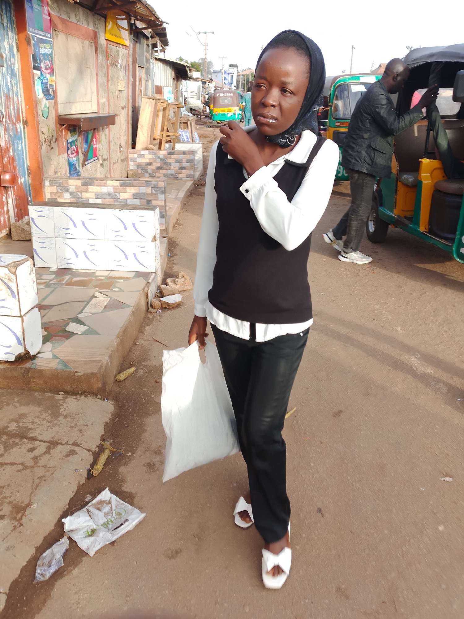 A young Black woman wearing a black headscarf, a white collared shirt, and black pants walks down a paved street in Gura Top, Nigeria, holding a white plastic bag. To her left are several market stalls with corrugated metal roofs and weathered wooden and metal structures. The stalls have tiled fronts and some have posters and signs displayed. The street is a mix of asphalt and dirt. Behind the woman, a man in a dark jacket and jeans is entering a green and yellow auto-rickshaw. More auto-rickshaws are visible in the background. The ground in front of the stalls is decorated with mosaic tiles. A piece of corn is lying on the ground in front of the woman, along with some discarded white plastic bags. The lighting suggests it is daytime.