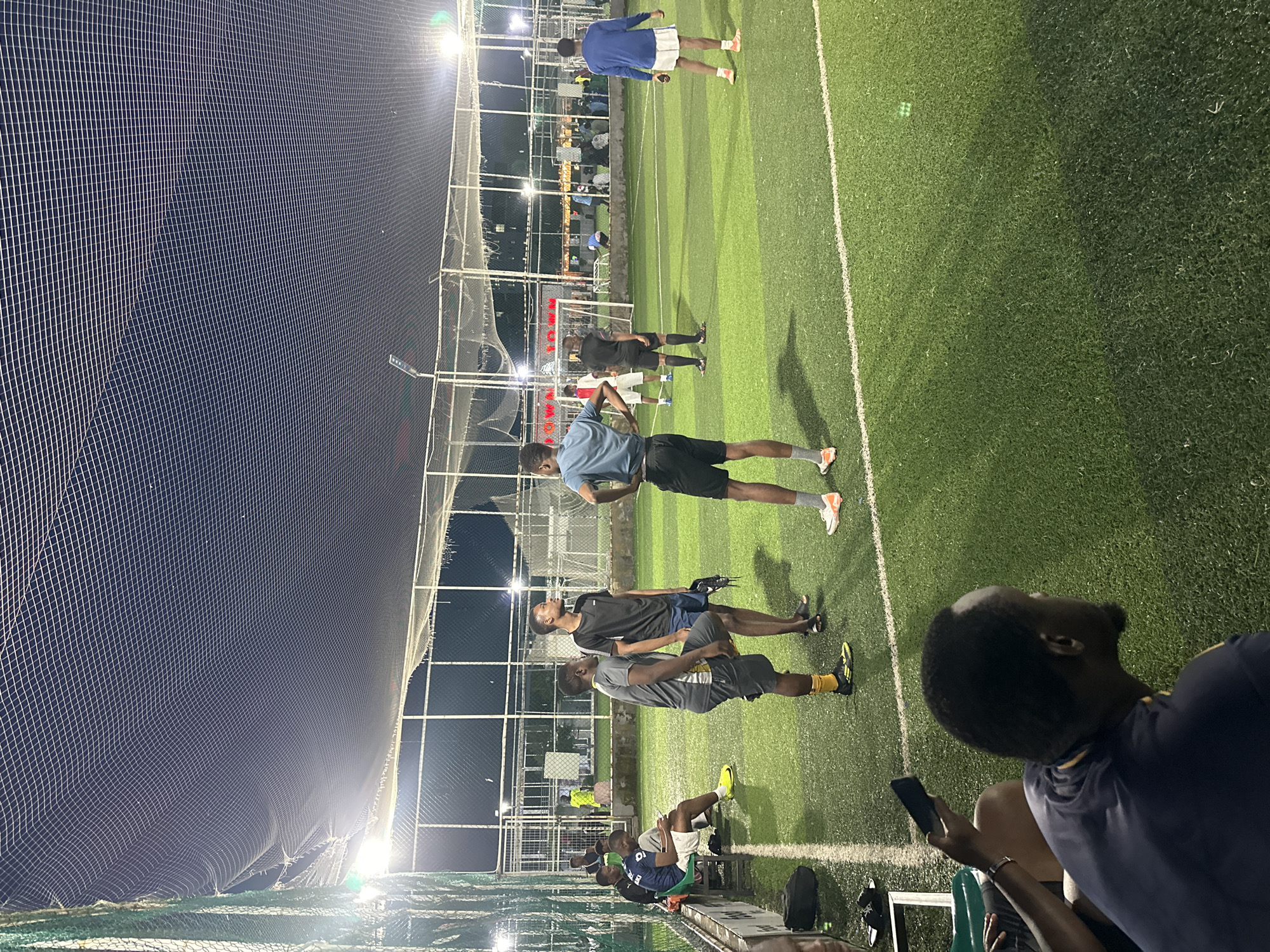 The image depicts an outdoor, artificial turf football pitch at night in Abuja, Nigeria. The pitch is illuminated by artificial lights, and a large net stretches overhead, enclosing the playing area. Several individuals are present on the pitch, some of whom appear to be playing or preparing to play football.
In the foreground, a man wearing a dark blue and yellow football jersey is seated, looking away from the camera. To his left, another person, partially visible, holds a dark smartphone.
Further into the pitch, a group of men are gathered. One man in a grey t-shirt and black shorts stands with his hands on his hips, facing left. Another man, in a black t-shirt and dark shorts, stands nearby. To his left, a man in a light blue t-shirt and black shorts is looking upwards. In the mid-ground, more individuals are seen scattered across the pitch, with one man in a blue long-sleeved shirt and white shorts walking towards the right of the frame.
In the background, a goalpost is visible, and other structures and individuals can be seen beyond the fence surrounding the pitch, suggesting a public or recreational facility. The overall scene is casual and active, indicative of a sports gathering.