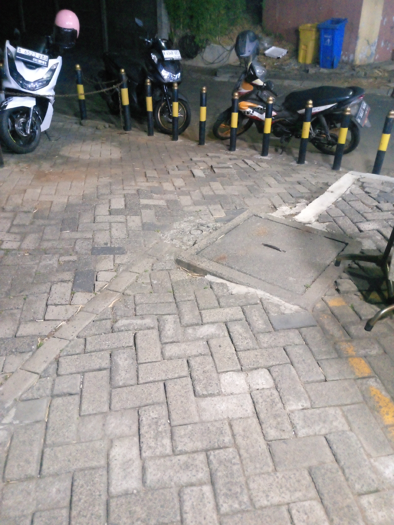 The image shows a street scene in Bitung Jaya, Indonesia, at night. Two motorcycles are parked on a brick-paved sidewalk. The motorcycle on the left is white and has a pink helmet on its handlebars. The motorcycle on the right is black and yellow. A manhole cover is visible in the foreground, and two trash cans, one yellow and one blue, are in the background. The street is lit by artificial lights, casting long shadows.