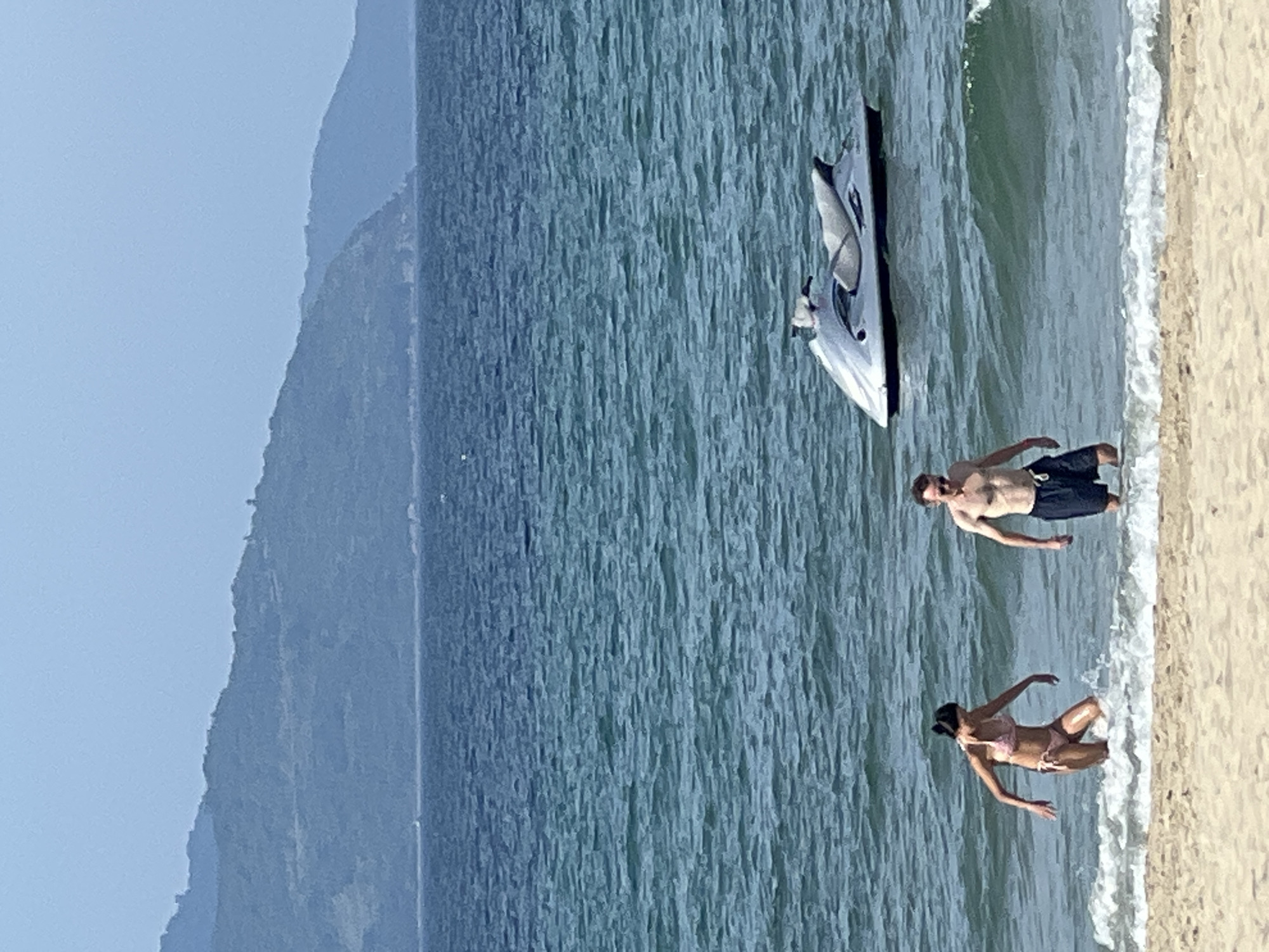 The image depicts a bright, sunny day at a beach, likely in Đà Nẵng, Vietnam, with a coastline featuring rolling, green hills in the background. The sea is a deep blue, with gentle waves lapping at the shore. In the foreground, a sandy beach leads into the water.
Two individuals are partially submerged in the shallow water. On the left, a woman wearing a pink bikini is facing away from the camera, with her right arm extended towards the water. On the right, a man in dark swim trunks is facing the camera, looking to his right.
Further out in the water, a silver jet ski is visible, bobbing gently on the surface. The overall scene suggests a leisurely day at the beach with recreational activities.