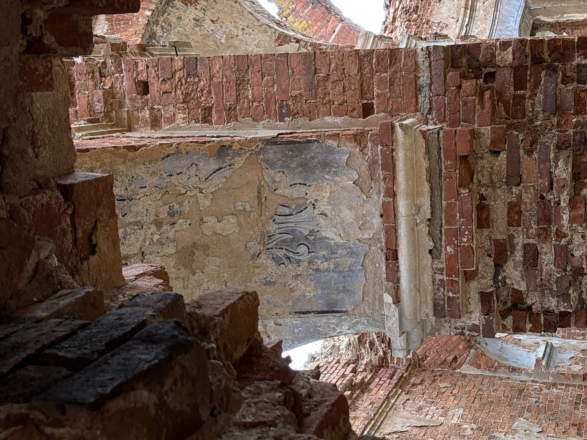 The image depicts the interior ruins of a building in Staroe Rakomo, Russia. The structure is primarily constructed of red brick, with significant sections decayed and crumbling. Plaster is visible on some wall surfaces, displaying remnants of decorative patterns in dark gray or black. The view is framed by sections of broken brick in the foreground, drawing the eye to the open spaces of the ruined structure. Light filters through gaps in the walls and ceiling.