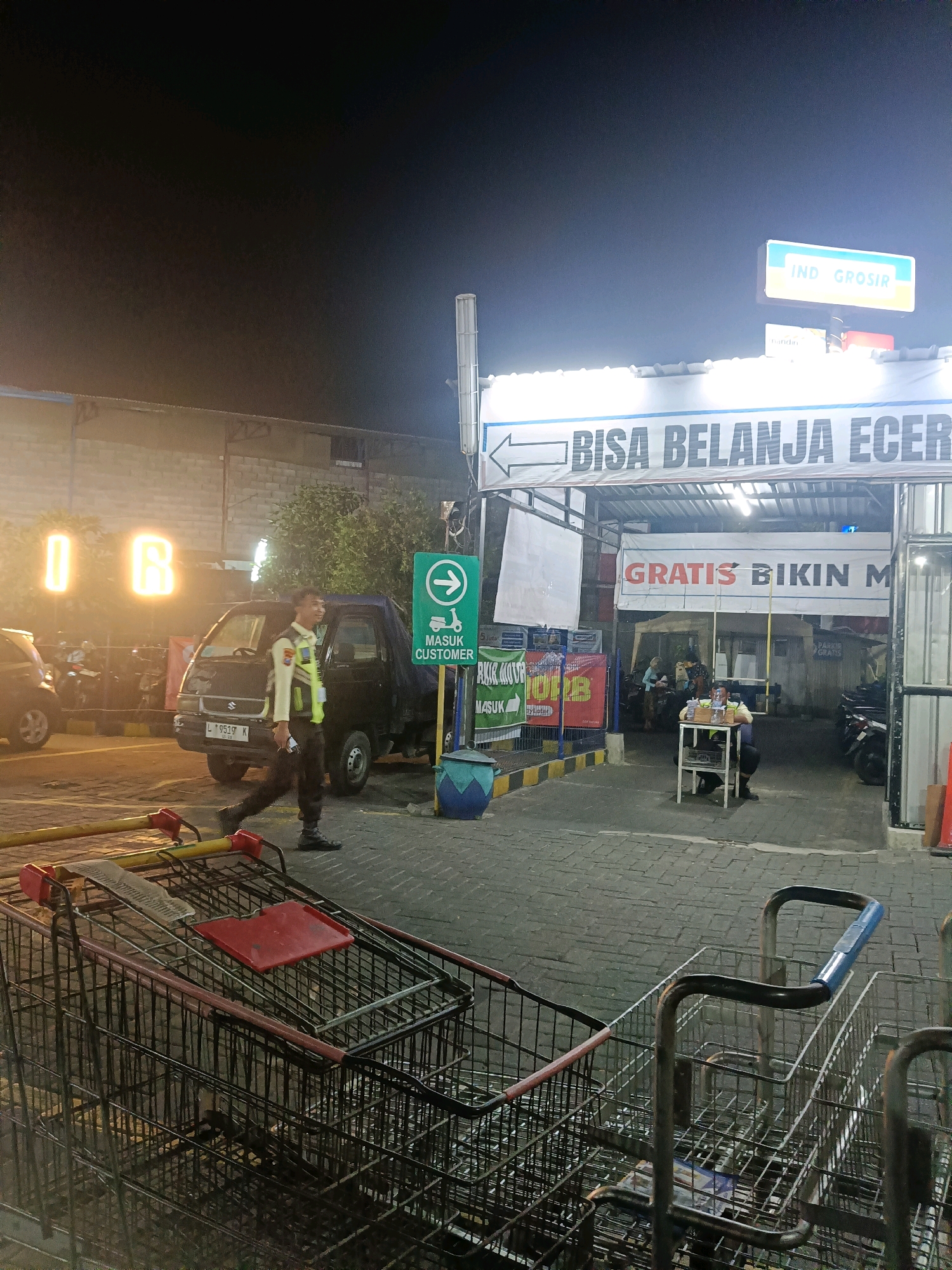 The image depicts the entrance of an Indogrosir store in Surabaya, Indonesia, at night. The store's signage is visible at the top, with "IND GROSIR" illuminated. A large banner across the entrance reads "BISA BELANJA ECER" which translates to "Can Shop Retail". Another banner below it says "GRATIS BIKIN M" likely indicating a free service.
In the foreground, several shopping carts are stacked, indicating a retail environment. A security guard in uniform, wearing a bright yellow vest over a dark uniform and pants, is walking towards the right of the frame. He appears to be holding a mobile phone. Behind him, a dark-colored pickup truck is parked, with a license plate visible that reads "L 9519 K".
Further into the store's entrance, a man is seated behind a small table, possibly another staff member or security personnel. He is also wearing a yellow vest. Several motorcycles are parked in the background, suggesting customer vehicles. The ground is paved with interlocking blocks. The overall atmosphere is that of a typical retail establishment at closing or during a less busy period at night. The lighting is artificial, with the store's lights casting a bright glow.