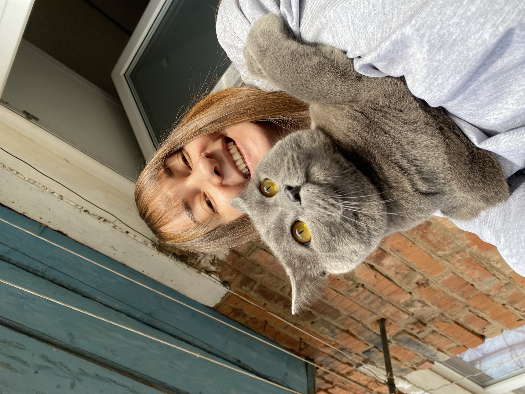 A woman and a gray cat are visible. The woman is smiling and has brown hair. The cat has yellow eyes. They are indoors, near a window and a wall made of blue wooden planks and red bricks. A clothesline is attached. The location is likely Votkinsk, Russia.