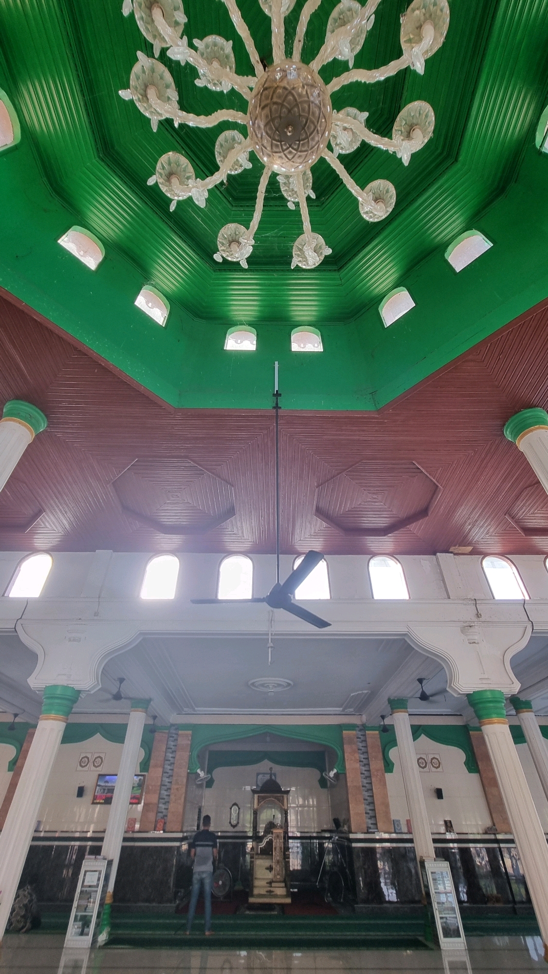 An interior shot of a mosque in Blangrambong, Indonesia, captures a low-angle view of a green and brown patterned ceiling. A large, ornate chandelier with clear glass elements hangs from the center of the ceiling, suspended by thin wires. Below, a dark ceiling fan is positioned directly under the chandelier. The ceiling transitions from a green upper section with arched windows to a brown, geometrically patterned section. Further down, white columns with green accents frame the view of the mosque's interior. Several arched windows with a bright light source are visible above the lower section of the mosque. In the foreground, a man stands facing away from the camera, towards an ornate wooden structure that appears to be a pulpit or minbar. Another person is partially visible to the right, and bicycles are parked along the back wall. The floor is covered in green carpet, common in mosques.