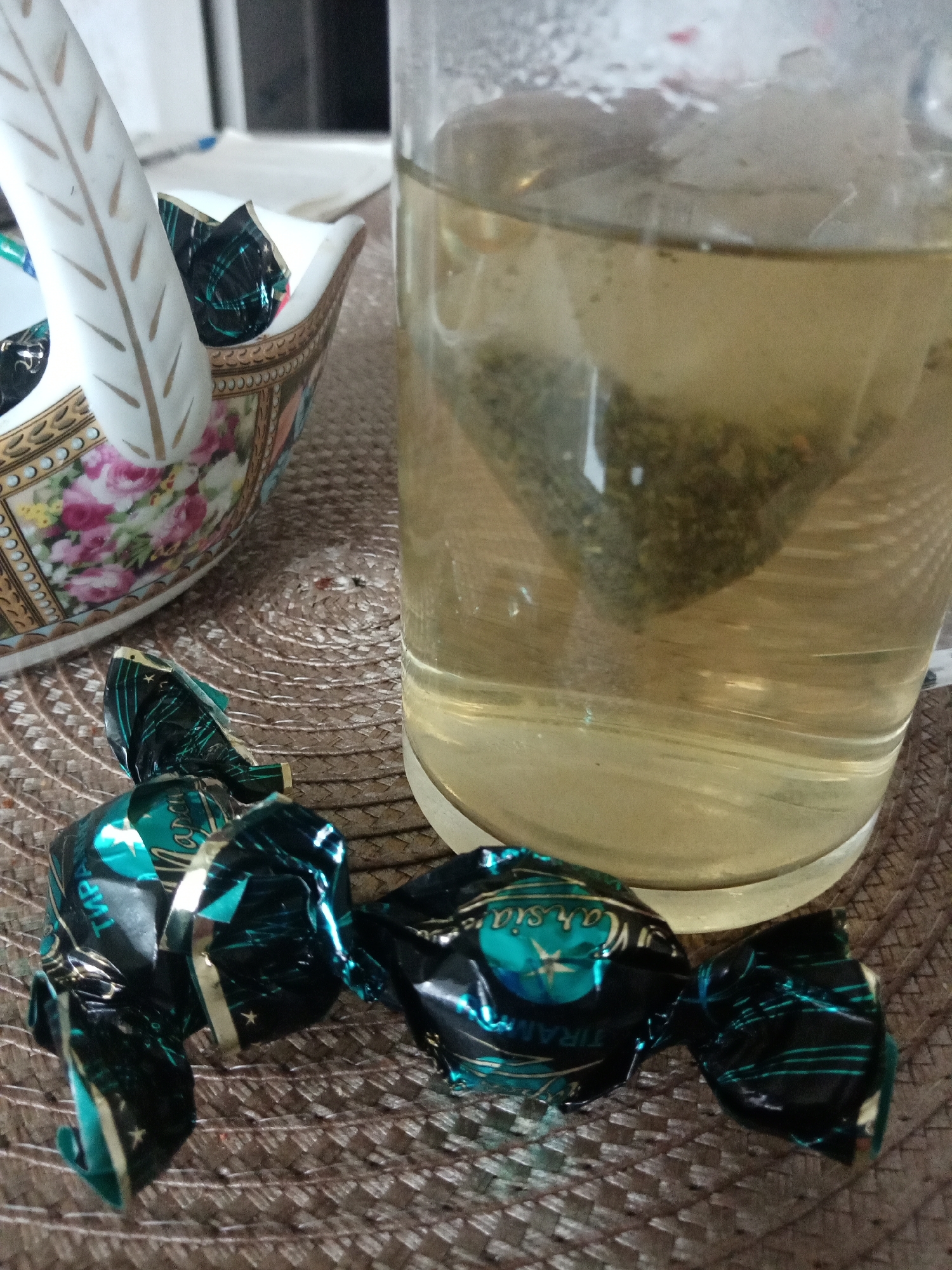 The image shows a table setting with a cup of tea, a porcelain candy dish, and three wrapped candies. The tea is a light color and has a tea bag steeping inside. The candy dish is decorated with floral patterns and gold accents. The candies are wrapped in blue and black foil, with the word "Tiramisu" visible on one. The table mat is woven in a circular pattern. The location in Elektrostal, Russia is unknown and not identifiable from the image.