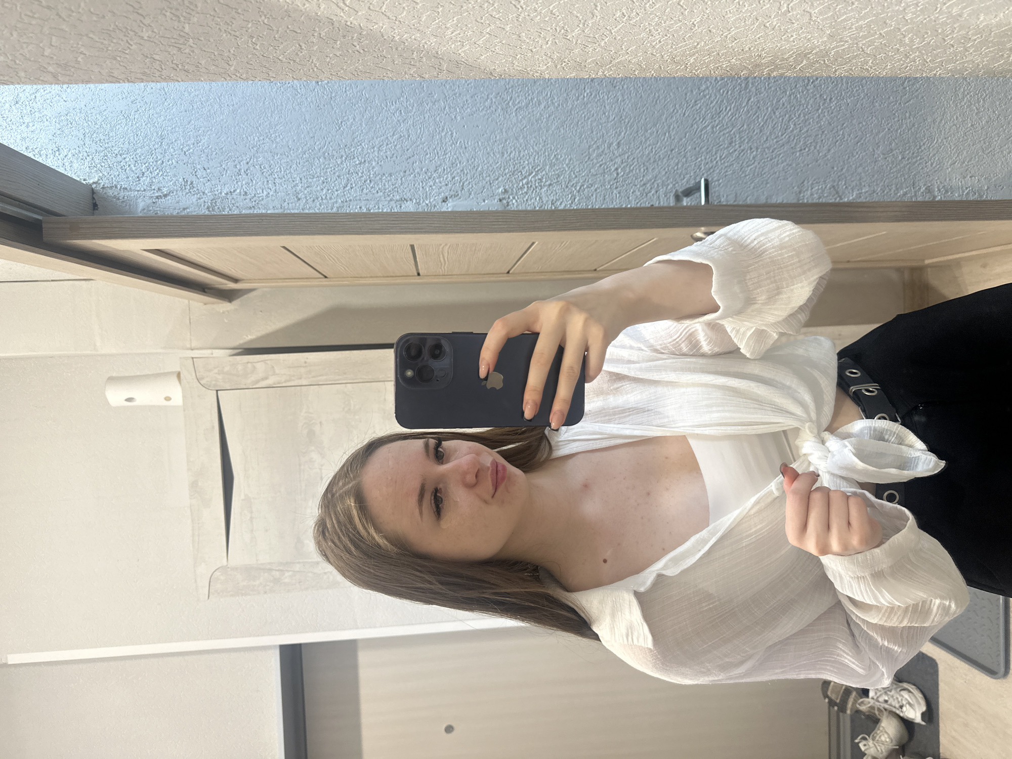 In an indoor setting, a young woman with long, brown hair stands in front of a mirror, taking a selfie with her phone. She is wearing a white, sheer button-down shirt tied at the waist over a white crop top, and black pants. The background includes a light-colored wall with a textured finish, a wooden sliding door, and a cabinet. To the left of the woman, a pair of white sneakers is visible on a gray mat. The lighting suggests an indoor environment, likely a home or dressing area.