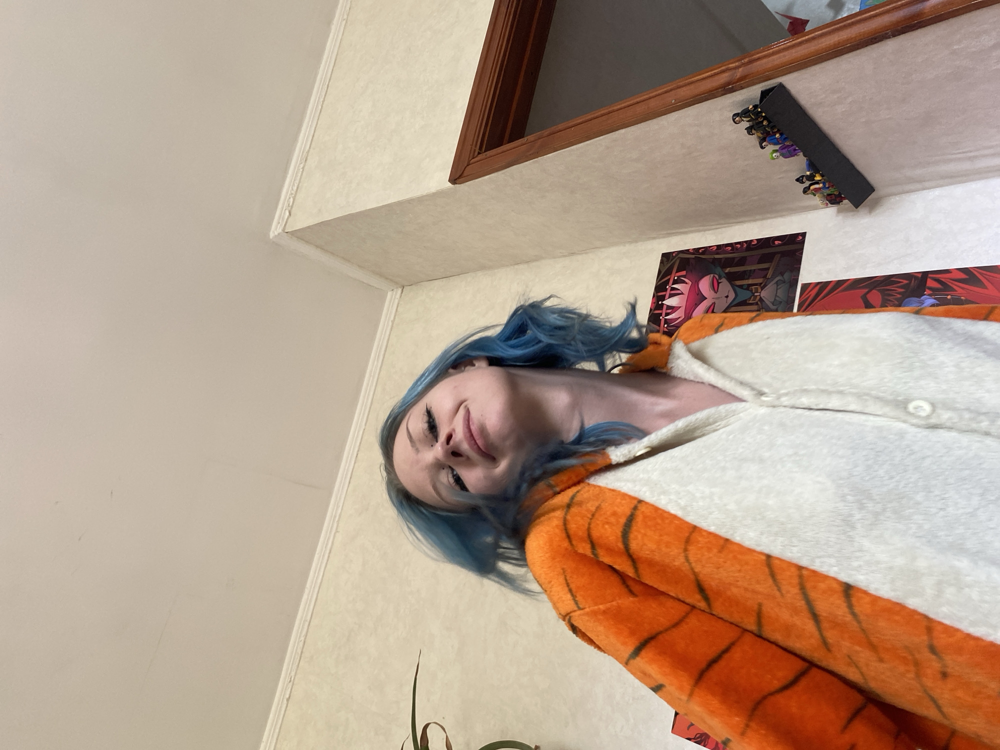 A person with blue hair is wearing an orange and white tiger-striped onesie. The person is looking down at the camera with a slight smile and their eyes are partially closed. The background appears to be a room with textured, light-colored walls and a wooden trim along the top. On the wall, there are posters with colorful illustrations. Above the posters, a black shelf is mounted, holding several small figurines. The lighting suggests an indoor setting, likely during the daytime. The overall mood seems casual and playful.