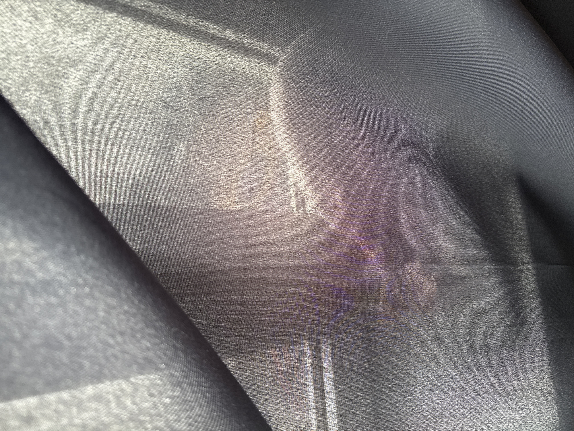 The image presents a close-up, abstract view dominated by a dark grey or black sheer fabric, likely a curtain or drape. The material has a fine, woven texture, visible as a subtle grid pattern, particularly where light interacts with it.
Light filters through the translucent fabric from an unseen source, illuminating various sections. This illumination creates areas of lighter grey, almost white, and reveals faint, indistinct shapes and color variations behind the material. In the central and right portions of the image, diffuse yellowish-brown and purplish hues are apparent, suggesting reflections or objects positioned beyond the curtain. These background elements are too blurred and obscured by the fabric's sheerness to be clearly identified, appearing as ethereal blurs and soft outlines. Some faint vertical and horizontal lines might suggest structural elements like window frames or furniture, but these remain highly ambiguous.
The scene conveys a sense of diffused light, indicating it is likely daytime. There are no identifiable people, animals, or specific activities taking place. The texture of the fabric and the interplay of light and shadow are the primary focus. No visible text is present in the image. While the location is provided as Khimki, Russia, there are no visual cues within the image itself to confirm this geographical context.