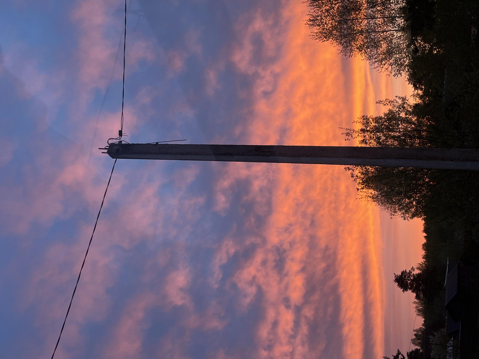 The image captures a vertical view of a sunset scene in Раздолье, Russia. An aged utility pole stands prominently in the foreground, with wires extending towards the sky. The sky exhibits a gradient of colors, ranging from orange and pink hues near the horizon to darker blue and purple tones higher up, indicating sunset. Trees and vegetation are visible in the upper-right section, silhouetted against the bright sky. Darker rooftops of buildings appear along the bottom-right corner, suggesting a residential or semi-urban area.