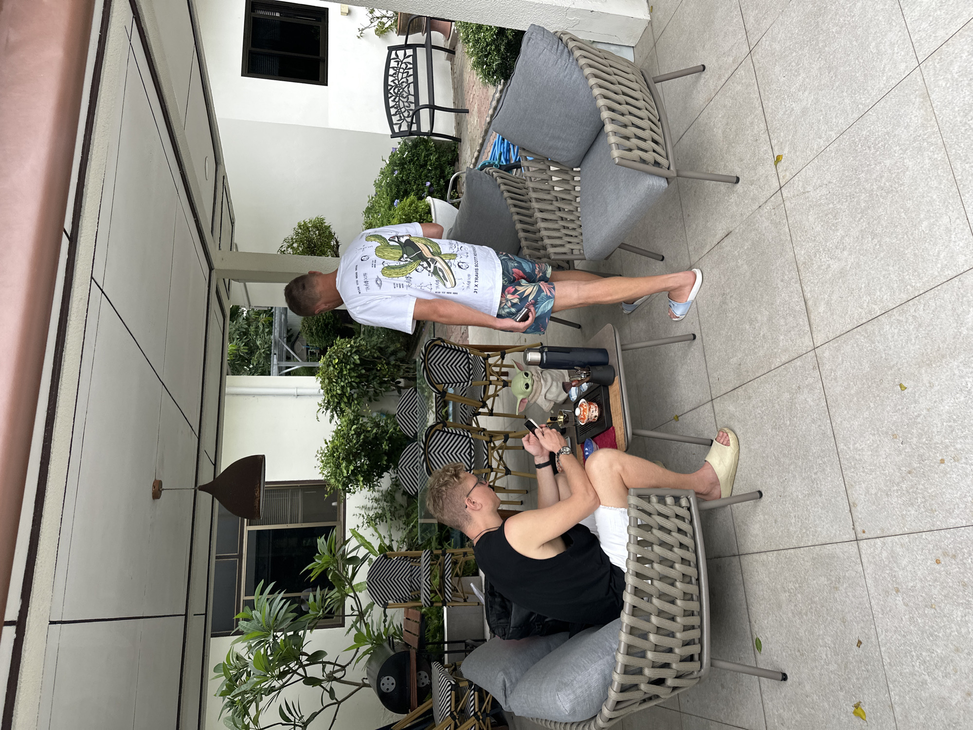 Two young men are on an outdoor patio. One man, wearing a black tank top and white shorts, is seated in a rattan chair and looking at his phone. In front of him is a small table with a cup, a figurine of Baby Yoda, and a bottle. The second man, wearing a white t-shirt with a graphic print and floral shorts, is standing and looking towards the seated man. The patio has a tiled floor and is covered by a roof. In the background, there are several chairs and a table, as well as plants and a wall of a building with windows.