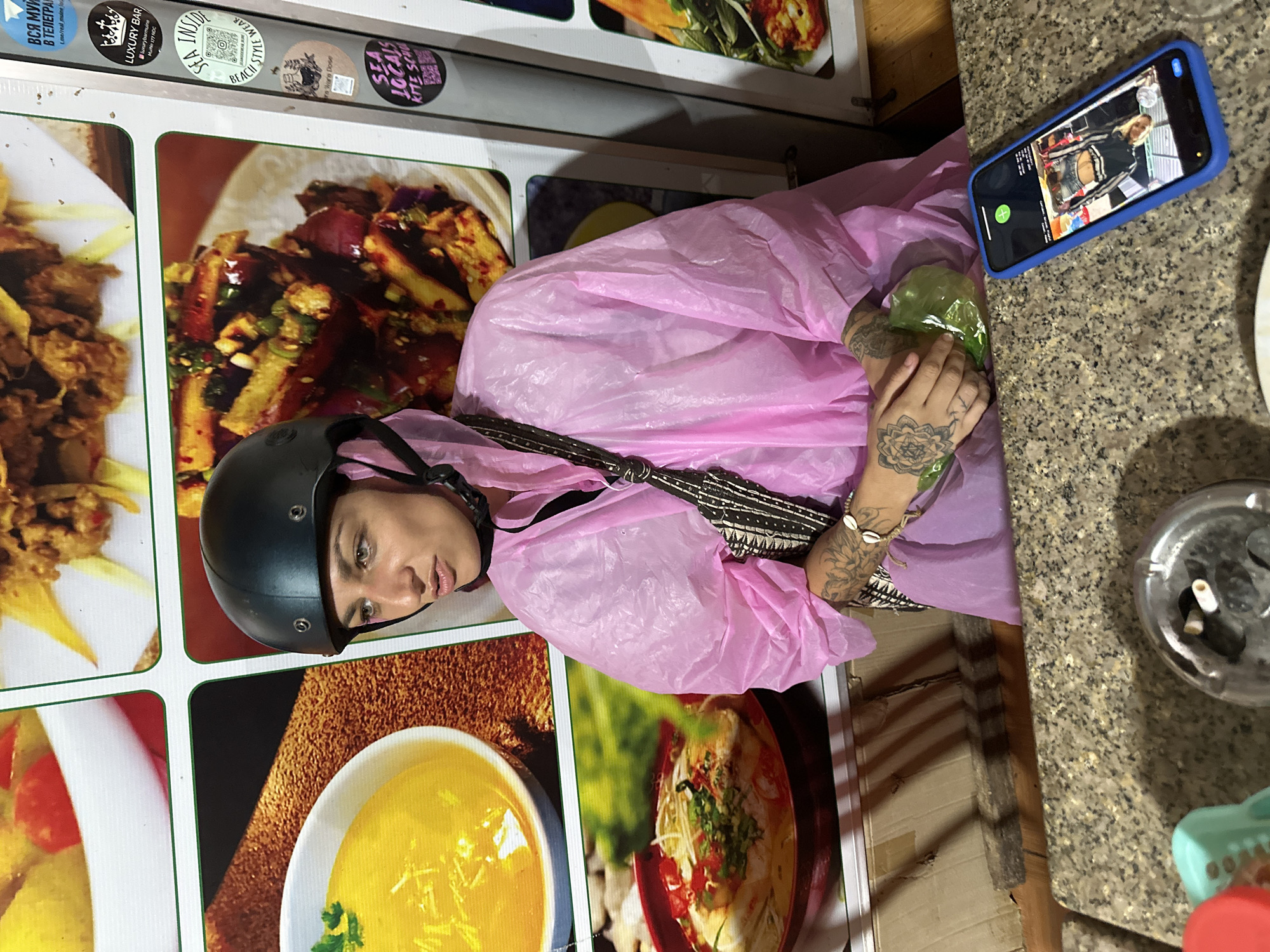 A young woman, wearing a black helmet and a bright pink plastic poncho, is captured in what appears to be an indoor or covered outdoor eatery setting. She has prominent tattoos on her forearms and hands, including a large mandala-like design on her left hand, and holds a small green object in her hands. She looks directly at the viewer with a neutral expression.
To her right, a smartphone in a blue case lies on a speckled granite-patterned counter. The phone's screen displays an image or video call of another person, a blonde woman dressed in a black and white top. Adjacent to the phone is a glass ashtray containing several cigarette butts.
The background consists of a wall adorned with large, vibrant pictures of various dishes, resembling a menu. Above these food images, several circular stickers provide details about local businesses. Visible text includes "ВСЯ МУЙ В ТЕЛЕГРАМ" (All Muy in Telegram), "LUXURY BAR" with additional contact information like "luxurybarmuine" and "MuNe 177 NDC", "SEA INSIDE" with "BEACH STYLE WEAR" and a QR code, and "SEA LOCALS KITE SCHOOL". Another partially visible sticker reads "WARE DOSE".
The artificial lighting suggests the scene takes place during the evening or night. The presence of the poncho indicates potentially rainy or cool weather. The combination of food pictures, a bar advertisement, and a kite school suggests a tourist-oriented area, consistent with the location context of Phu Thuy Ward, Vietnam.