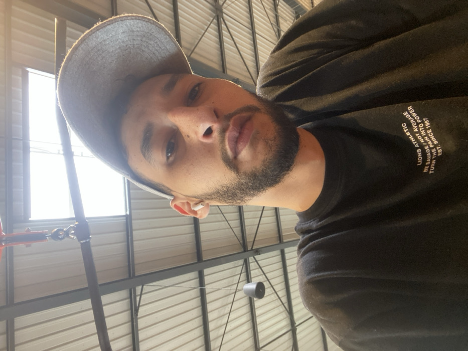 A close-up, upward-angled portrait shot depicts an adult male indoors in Hœnheim, France. The male, facing the camera directly, possesses light to medium skin, short dark hair partially covered by a backward-facing grey baseball cap, a dark beard and mustache, and visible freckles on his nose and cheeks. A white wireless earbud is present in his left ear. He wears a black t-shirt with white text reading "LIONS ATHLETIC," "BE SAVAGE / NOT AVERAGE," "TURN THE PAIN INTO POWER," and "EST. SINCE 1987." The background reveals an industrial-style ceiling structure featuring exposed dark metal beams and a light-colored corrugated roof. A large, bright skylight is prominent overhead, serving as the primary light source. A dark cylindrical light fixture hangs from the ceiling on the left, and a red metallic object, possibly part of an overhead mechanism, is partially visible in the upper left corner of the frame.