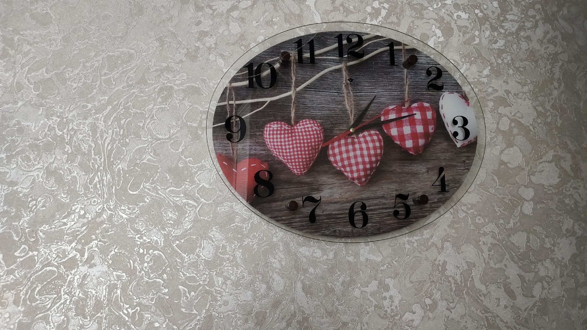 A round clock with a diameter of approximately 30 cm is displayed on a textured, light-colored wall. The clock face depicts a rustic wooden background with several decorative hearts hanging from a branch. The numbers on the clock are black and are positioned around the circumference. The hour hand is pointing at the number 11, and the minute hand is pointing at the number 3. The clock is framed by a thin black edge and a transparent outer layer. The wall behind the clock has a raised, abstract pattern with variations in light and shadow, creating a marbled effect.