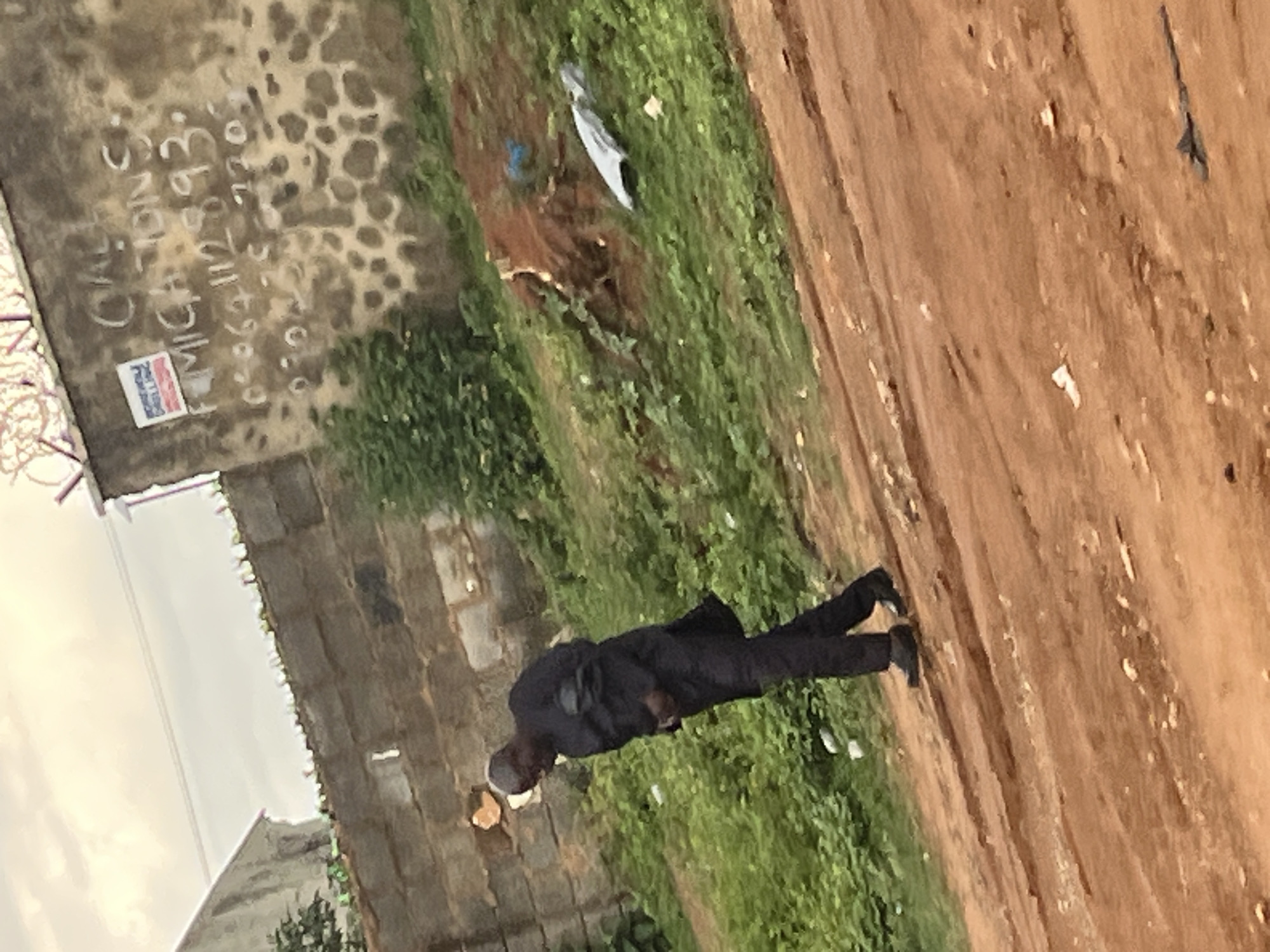 A medium shot of a dark-skinned man wearing a dark suit and light-colored skullcap is walking on a dirt path. The dirt path is to the right of the frame. To the left of the path, there is green grass and some garbage. A building made of gray bricks is behind the man, and graffiti and a sign are on the side of the building. The graffiti includes a telephone number. In the background is a cloudy sky and the roof of another building. The location is likely in Abuja, Nigeria, based on visual cues.