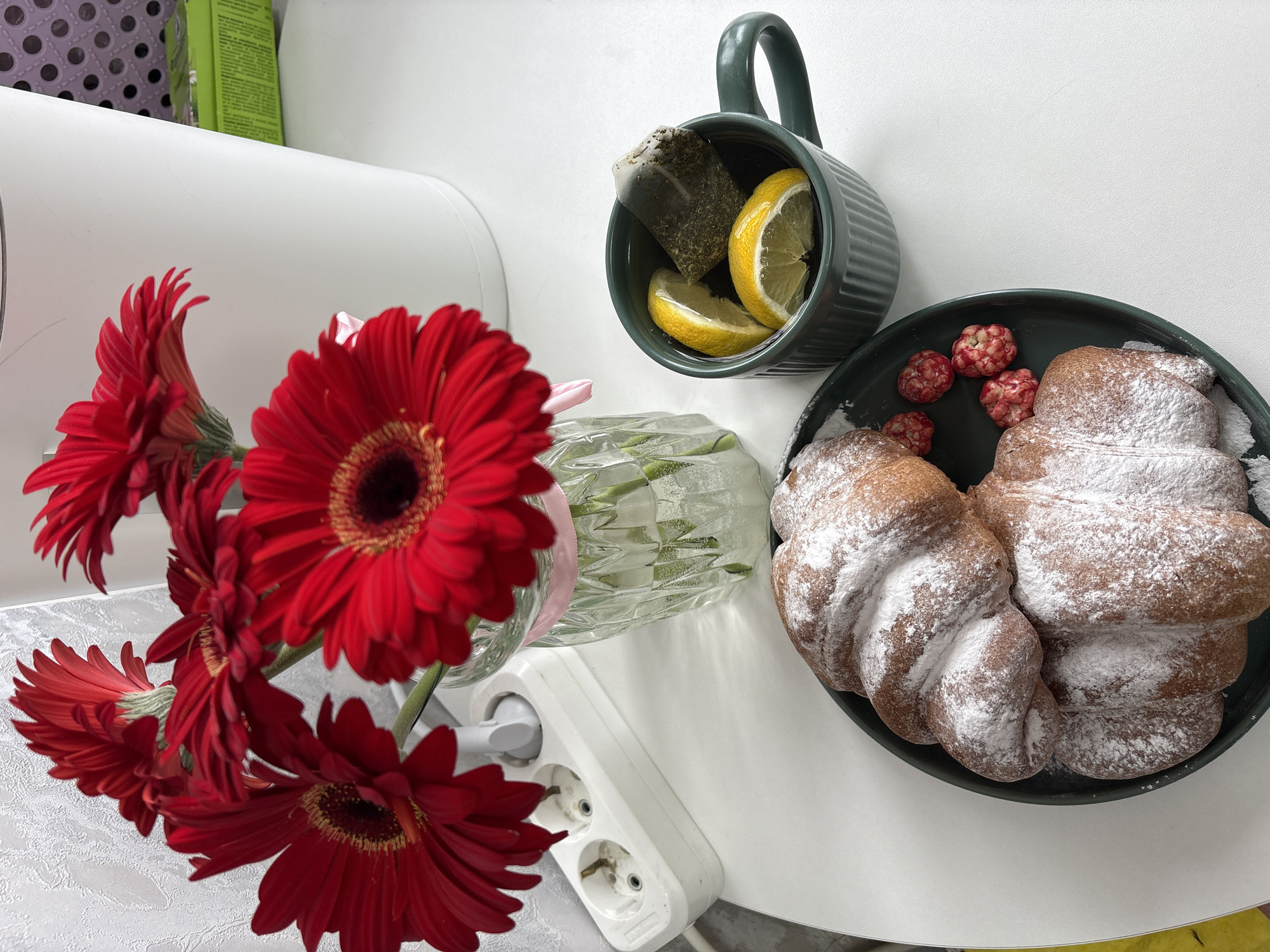 The image depicts a still life scene, likely indoors, on a white table. A vase containing red Gerbera daisies is placed to the left, partially obscuring a white power strip with a plug inserted. A green mug filled with tea, lemon slices, and a tea bag is positioned in the upper center. Adjacent to the mug, on the right, is a green plate containing two pastries dusted with powdered sugar and several small, red, berry-like candies. The setting suggests a domestic environment, and no specific details confirm the location in Chaikovsky, Russia.
