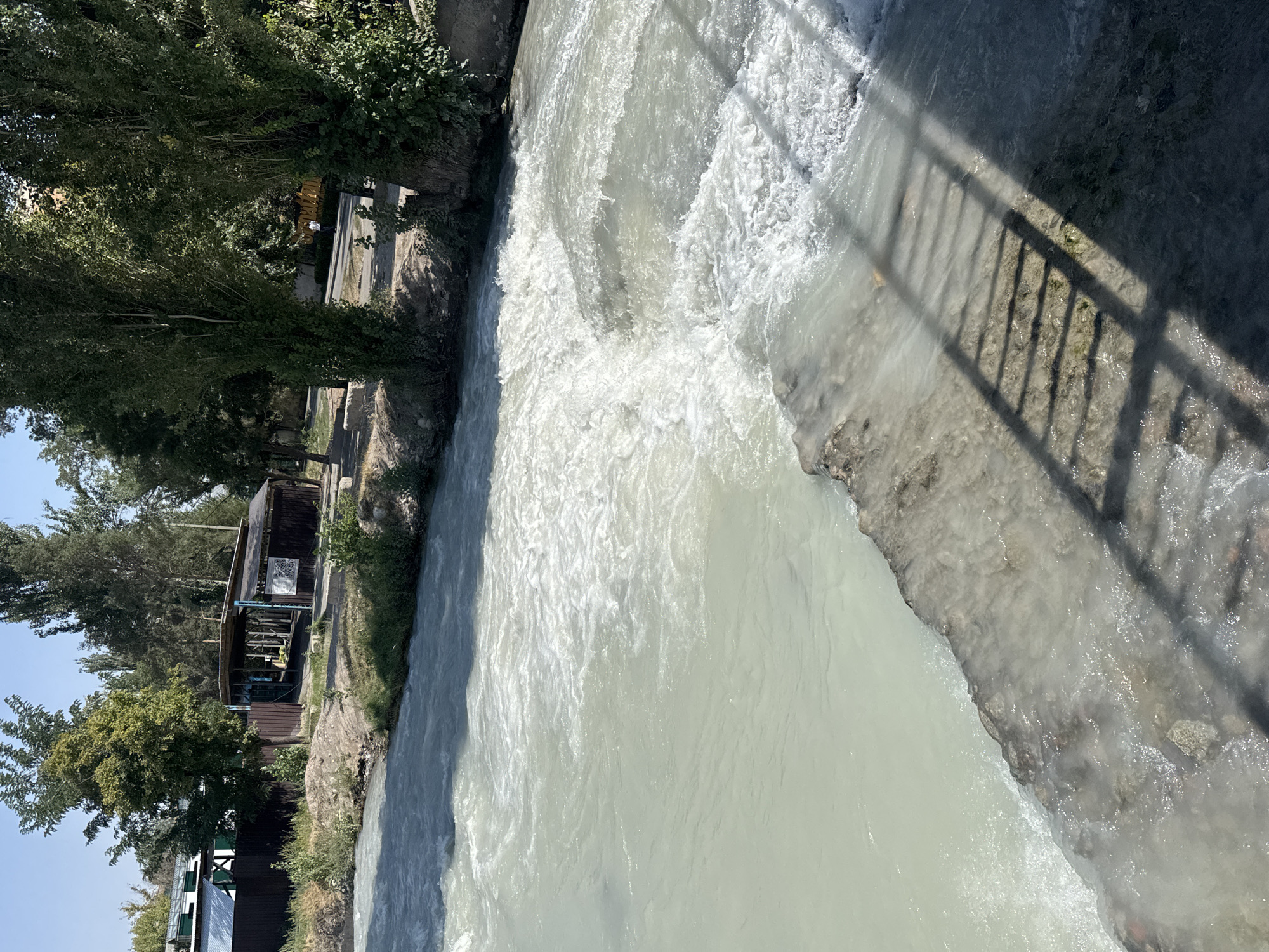 This image captures a view of the Ala-Archa River flowing through Bishkek, Kyrgyzstan. The river exhibits turbulent, whitish-gray water, suggesting a strong current and possibly sediment. On the left bank, there is a densely wooded area with lush green trees under a clear blue sky. Further back, a low-lying structure with a dark roof and brown siding is visible, bordered by a dark brown fence. The right bank of the river is marked by a concrete embankment, where the shadow of a railing is cast across the water's edge, hinting at a bridge or walkway overhead. The overall scene depicts a natural waterway within an urban or semi-urban environment.