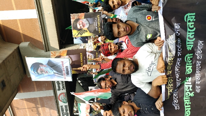 A daytime outdoor protest is depicted on a street in Dhaka, Bangladesh, against the backdrop of the Bangladesh Secretariat building and an elevated concrete structure overhead. A crowd of predominantly male individuals, primarily young to middle-aged, participates in the demonstration. They hold numerous signs, posters, and flags. A large black banner spanning the foreground displays Bengali text protesting the alleged assassination of "Ayatollah Ali Khamenei by America-Israel." Individual posters held aloft feature images of prominent figures, including Ayatollah Ali Khamenei, Ayatollah Ruhollah Khomeini, General Qasem Soleimani, and Sayyid Hassan Nasrallah. Several Iranian national flags and various green flags are visible among the crowd. Participants generally wear casual attire and exhibit serious, focused expressions.