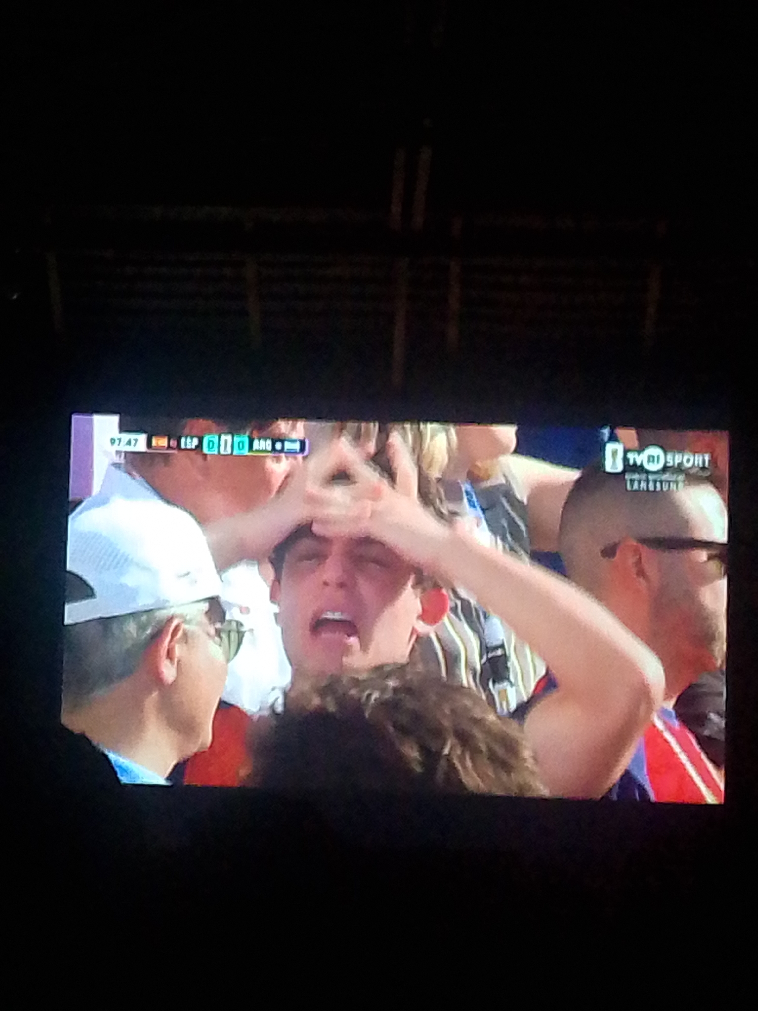 The image displays a screen, likely a television, showing a sports broadcast with a scoreboard overlay. The scoreboard indicates "97:47 ESP 010 ARG". A man in the foreground, with his mouth open, is the primary subject, with his face partially obscured by his hand which forms a triangle shape. To his left, another man is visible wearing a white cap and sunglasses. Behind them, other people are present in a crowd setting, some wearing jerseys. A logo for "TVRI SPORT" is visible in the upper right portion of the screen. The scene is dark, suggesting it is nighttime or the surrounding environment is dimly lit, with the screen being the main source of light. There is no discernible connection to the city of Meunasahmee, Indonesia.
