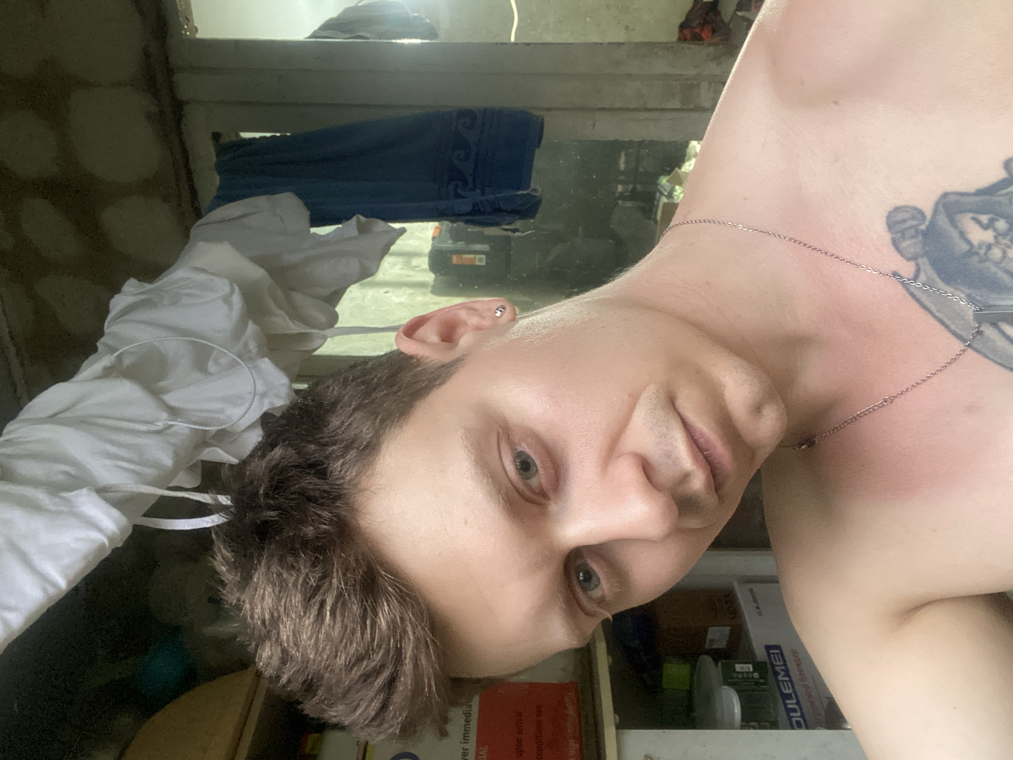 A shirtless man with short brown hair and blue eyes is looking directly at the camera. He is wearing a silver chain around his neck with a pendant. He has a tattoo on his chest. The background appears to be an outdoor setting, possibly a backyard or a rural area, with clothes hanging on a line and some containers in the background. The lighting suggests it is daytime. The image does not provide any specific visual cues that would allow for the identification of the location as Samara, Russia.