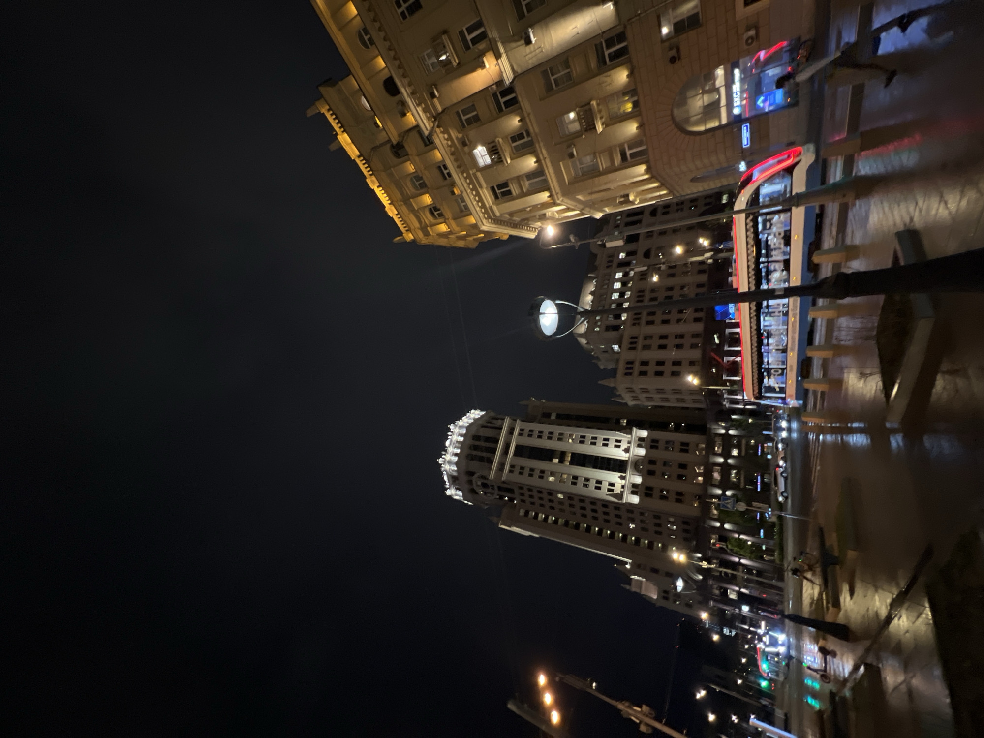 The image depicts a nighttime urban scene in an unknown city. Dominating the view are several tall buildings, illuminated with artificial light, contrasting against the dark sky. The buildings exhibit varying architectural styles, with one showcasing ornate details at its top. Streetlights punctuate the scene, reflecting off what appears to be wet pavement, suggesting a recent rain. The composition is tilted, offering a unique perspective of the city's vertical landscape. A blurred figure is discernible on the right side of the frame.
