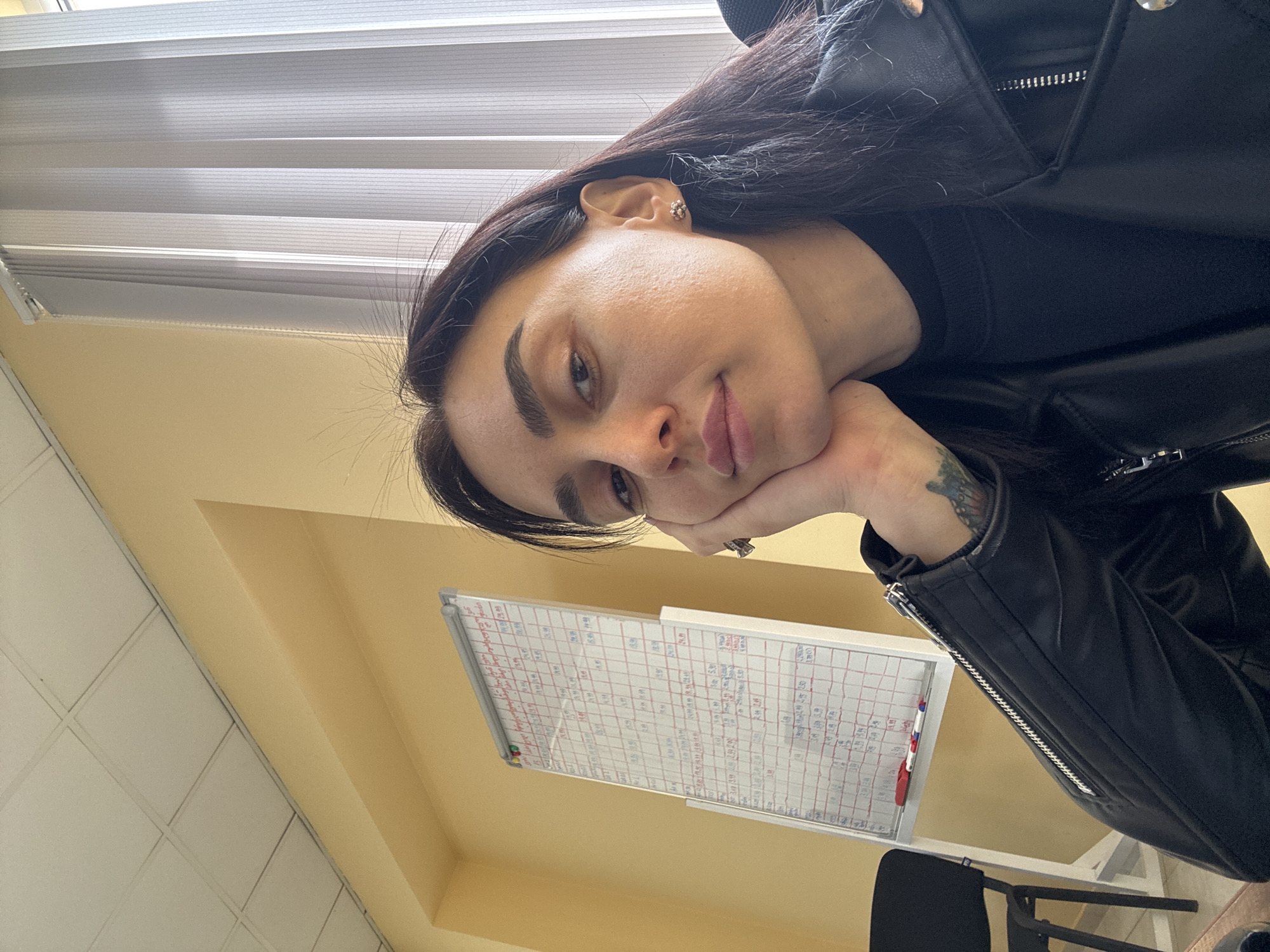 A woman with dark hair and a tattoo on her left forearm is resting her face on her hand. She's wearing a black leather jacket over a black top. Behind her, a whiteboard is filled with handwritten notes and grids, likely related to scheduling or data tracking. The background includes a wall and a partially visible window with blinds. A black chair is visible near the bottom of the frame. While the image provides interior details, there is insufficient information to determine the precise location in Tbilisi, Georgia.