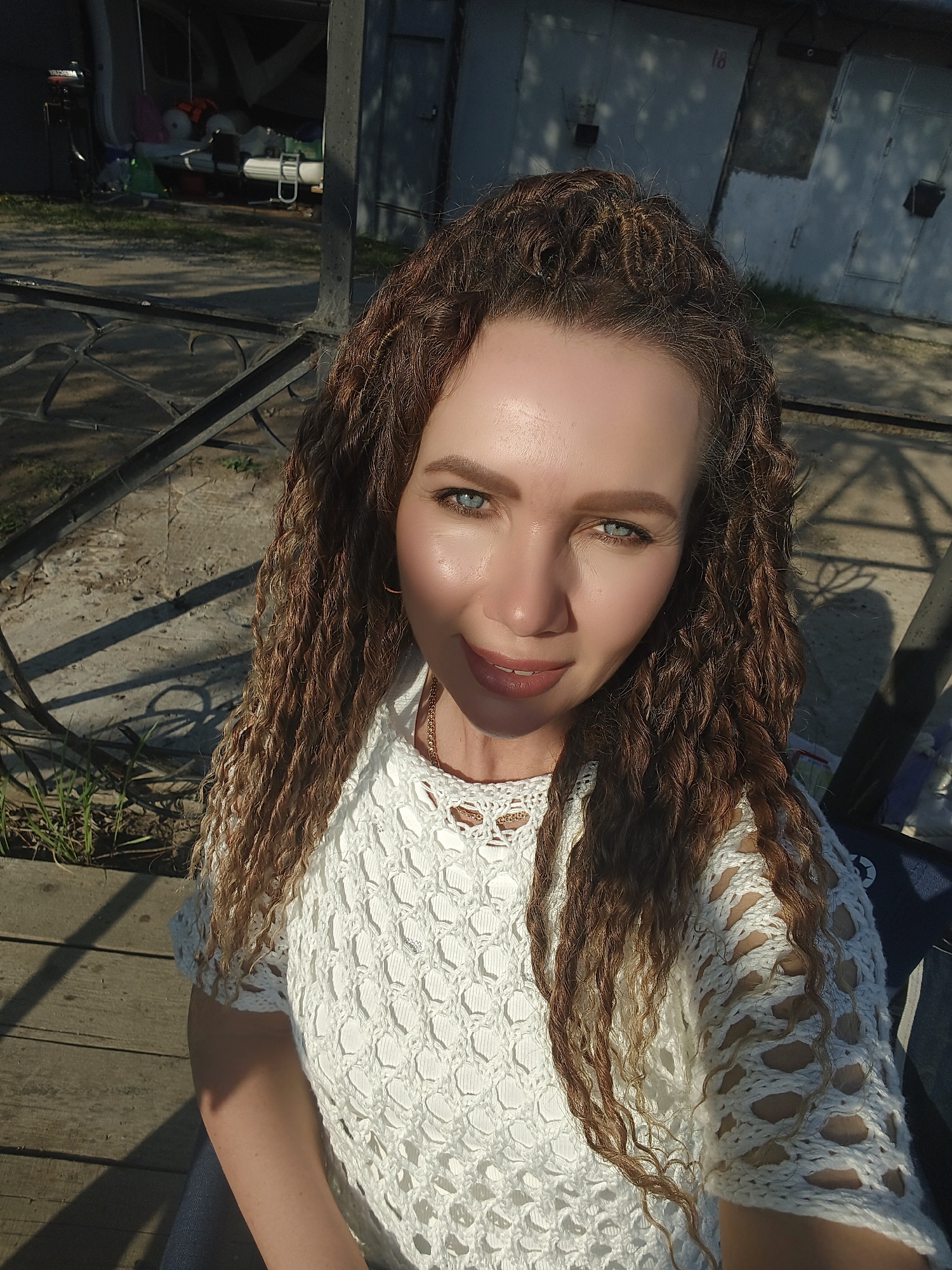 A woman with curly brown hair and blue eyes is in the city of Unknown, Unknown. She is wearing a white crochet-style top. The background includes a concrete surface with shadows, a black metal fence, and buildings with garage doors. The lighting suggests it is daytime.