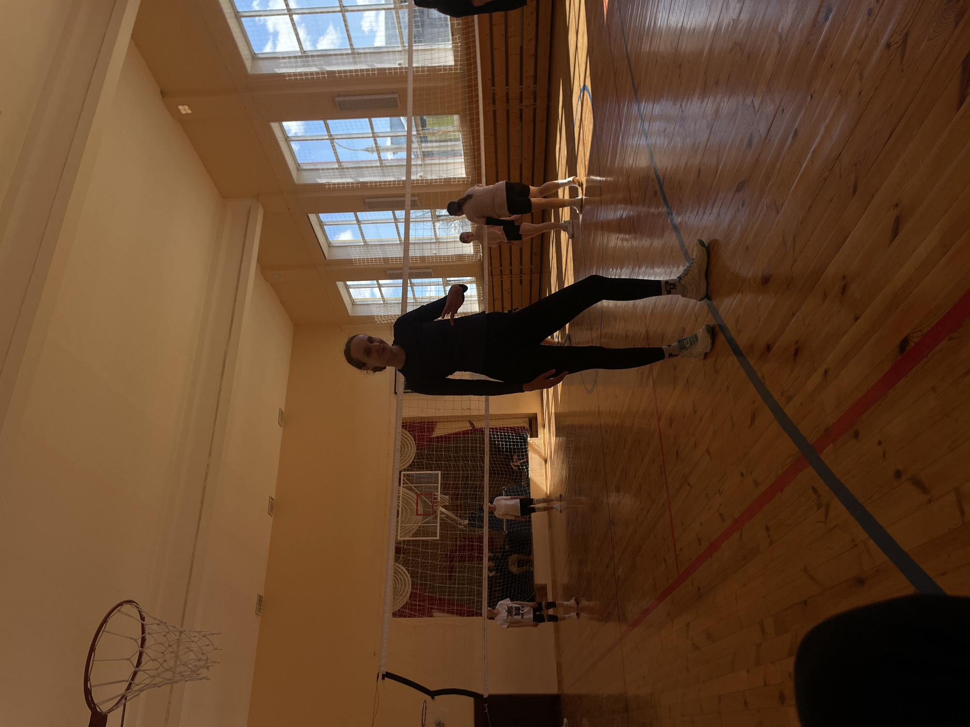 The image depicts the interior of a gymnasium. The location is Подшивалово, Russia. There are several people present. A woman in black athletic wear stands facing the camera. In the background, a few other individuals are engaged in volleyball practice near a net. Basketball hoops are visible on one end of the gym. The floor is wooden with visible lines. Natural light enters through windows in the ceiling.