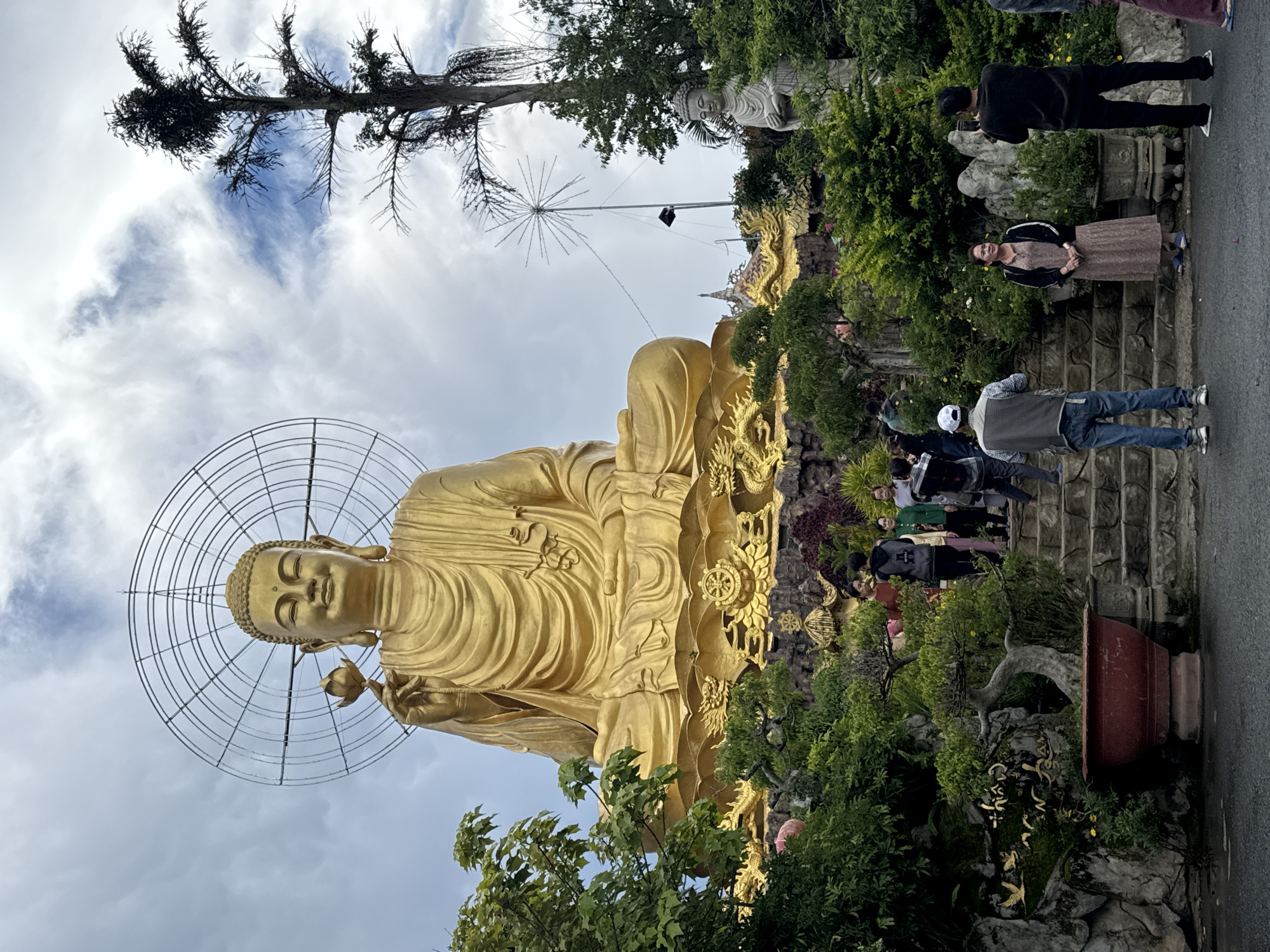 In the city of Lâm Viên - Đà Lạt, Vietnam, a large golden Buddha statue sits in meditation, holding a lotus flower. The statue is situated on a tiered, ornately decorated platform, adorned with golden dragons and Buddhist symbols. The surrounding environment is lush with greenery, featuring manicured trees and plants. Several people are visible, some standing on stone steps leading up to the platform, while others are taking photographs. A smaller, stone Buddha statue is also present among the foliage to the right of the main golden statue. The sky above is partly cloudy, with a large, circular metal structure behind the golden Buddha, possibly a halo or part of a light fixture. The scene appears to be a tourist or religious site.