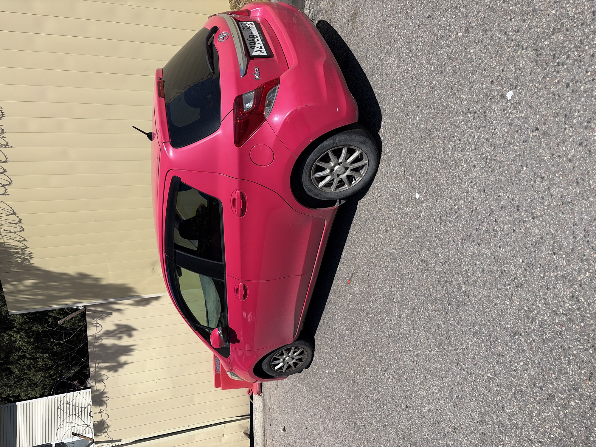 A bright fuchsia pink Toyota Yaris (identifiable by the "Vitz" badging and Toyota emblem) hatchback is parked on an asphalt surface, oriented facing left. The vehicle exhibits a Russian license plate "A296BH 323 RUS" on its rear, indicating its registration in Krasnodar Krai, Russia, which includes the city of Sochi. The car features dark tinted rear windows, a rear wiper, a roof-mounted antenna, and multi-spoke alloy wheels. Minor dirt is visible on the lower body panels, and a slight scuff or dent is present on the passenger-side rear bumper.
The asphalt pavement beneath the car is mottled grey with visible aggregate and contains scattered small debris. To the immediate left of the car's front end, a red rectangular barrier or object with faint graffiti is partially visible.
In the background, a tall, light beige-colored corrugated metal wall extends horizontally. Above this wall on the left, a white-slatted building or structure is visible, alongside dark green dense foliage. Barbed wire (razor wire) runs along the top of a fence structure in the upper left portion of the image. Strong sunlight from the upper right casts distinct shadows of what appear to be tree branches and the barbed wire onto the beige wall, and a shadow from the car is cast onto the asphalt to its left. No individuals or other active elements are present in the scene.
