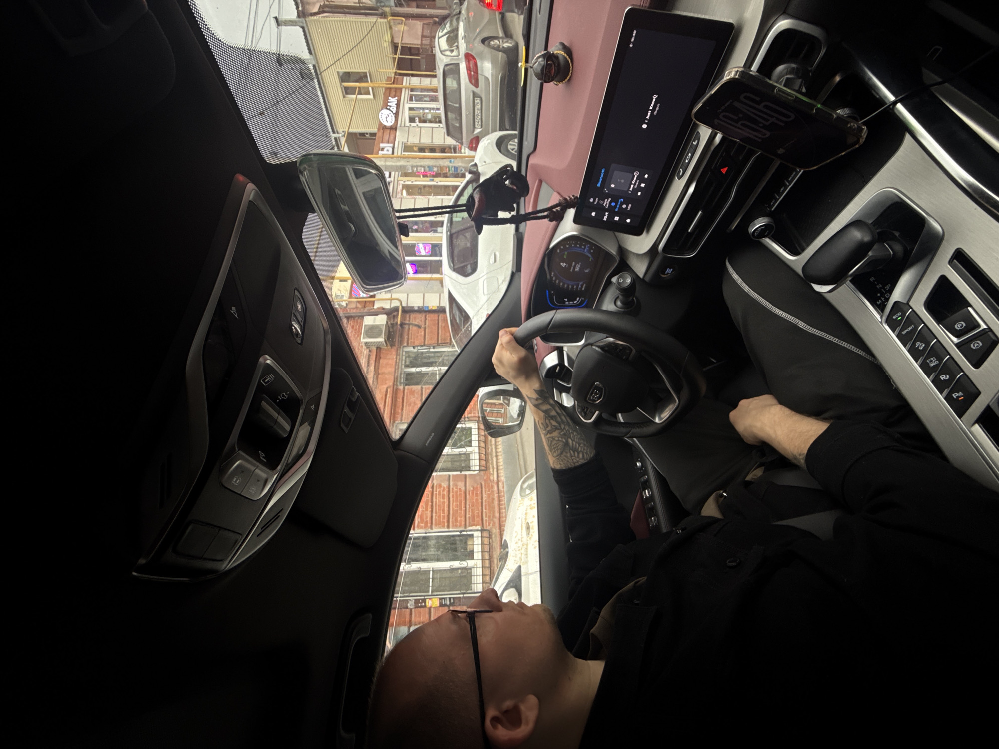 A person is driving a car in Rostov-on-Don, Russia. The view through the windshield shows a street with parked cars and buildings. The car's interior includes a dashboard with a screen, a steering wheel, and a phone mounted on the dashboard. The person driving is wearing glasses and a black jacket. The driver has a tattoo on their forearm.