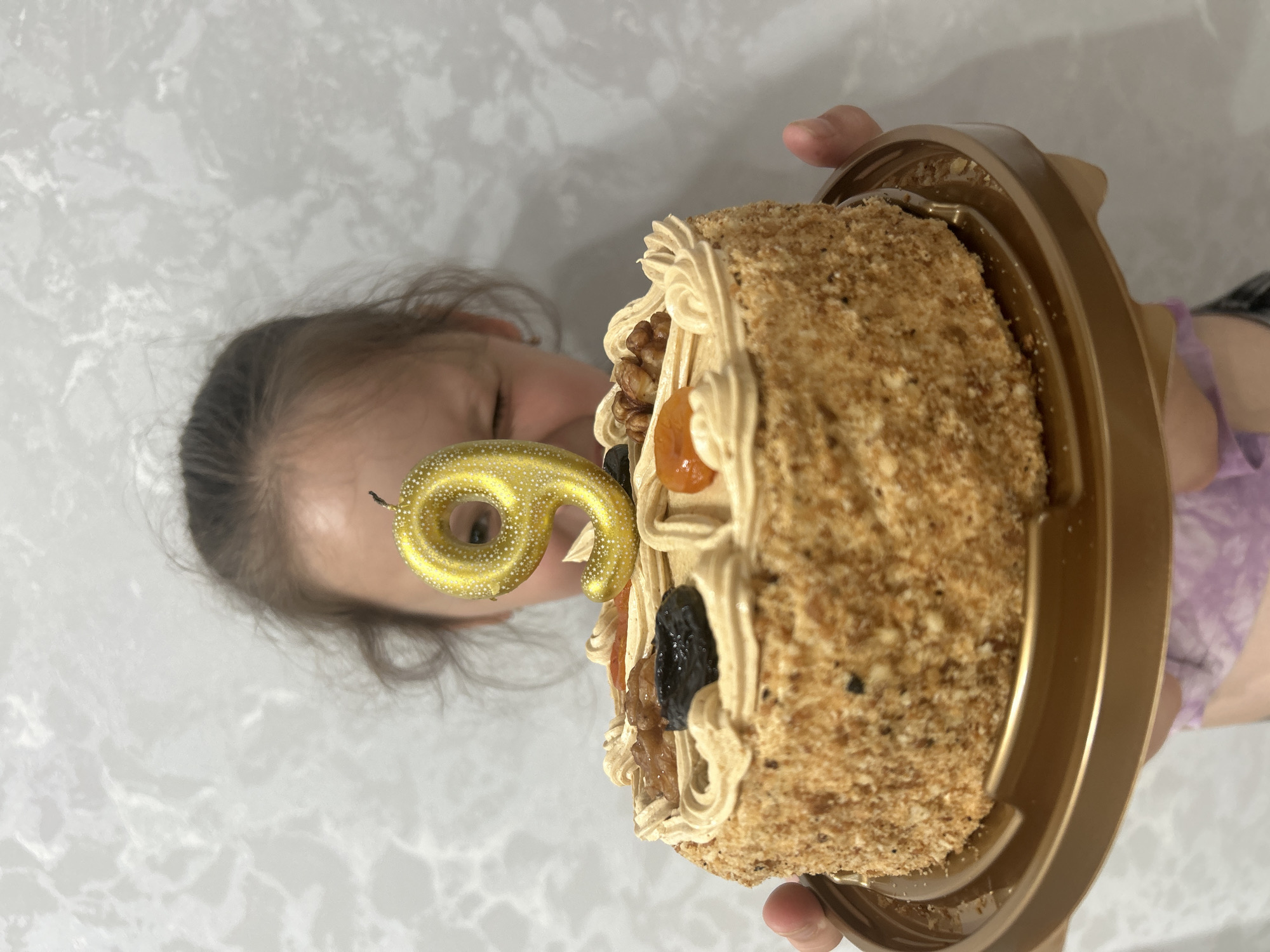 A child holds a cake with a lit number "9" candle in front of their face, obscuring most of their features. The cake is round and covered in a crumb coating, with decorative swirls of frosting and toppings of dried fruit and nuts. The background is a light-colored, textured wall, consistent with interior home decor. The image appears to be taken indoors, possibly in a domestic setting. There is no information in the image to suggest it is located in the city of Chaikovsky, Russia.