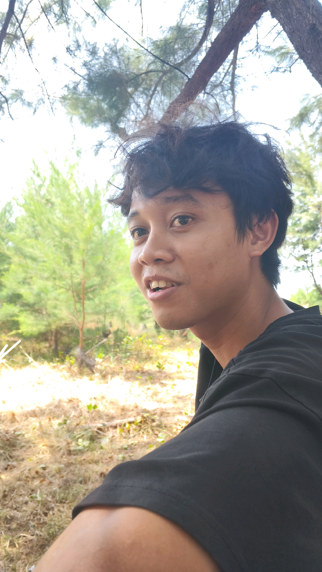 A close-up, outdoor, selfie-style photograph features a young East Asian male with messy black hair and medium-toned skin. He is wearing a black t-shirt and is looking slightly to the left of the camera, with his mouth slightly open and teeth visible. The background is blurred, showing greenery and dry foliage, indicative of a natural, possibly semi-arid environment. Above and behind him, tree branches and leaves are visible, suggesting he is outdoors under a canopy. The lighting appears to be natural daylight, with some areas appearing brighter due to sunlit foliage. The image composition is a portrait-style shot focused on the individual. While the image itself does not contain specific landmarks, the description states it is located in the city of Pabeanilir, Indonesia.