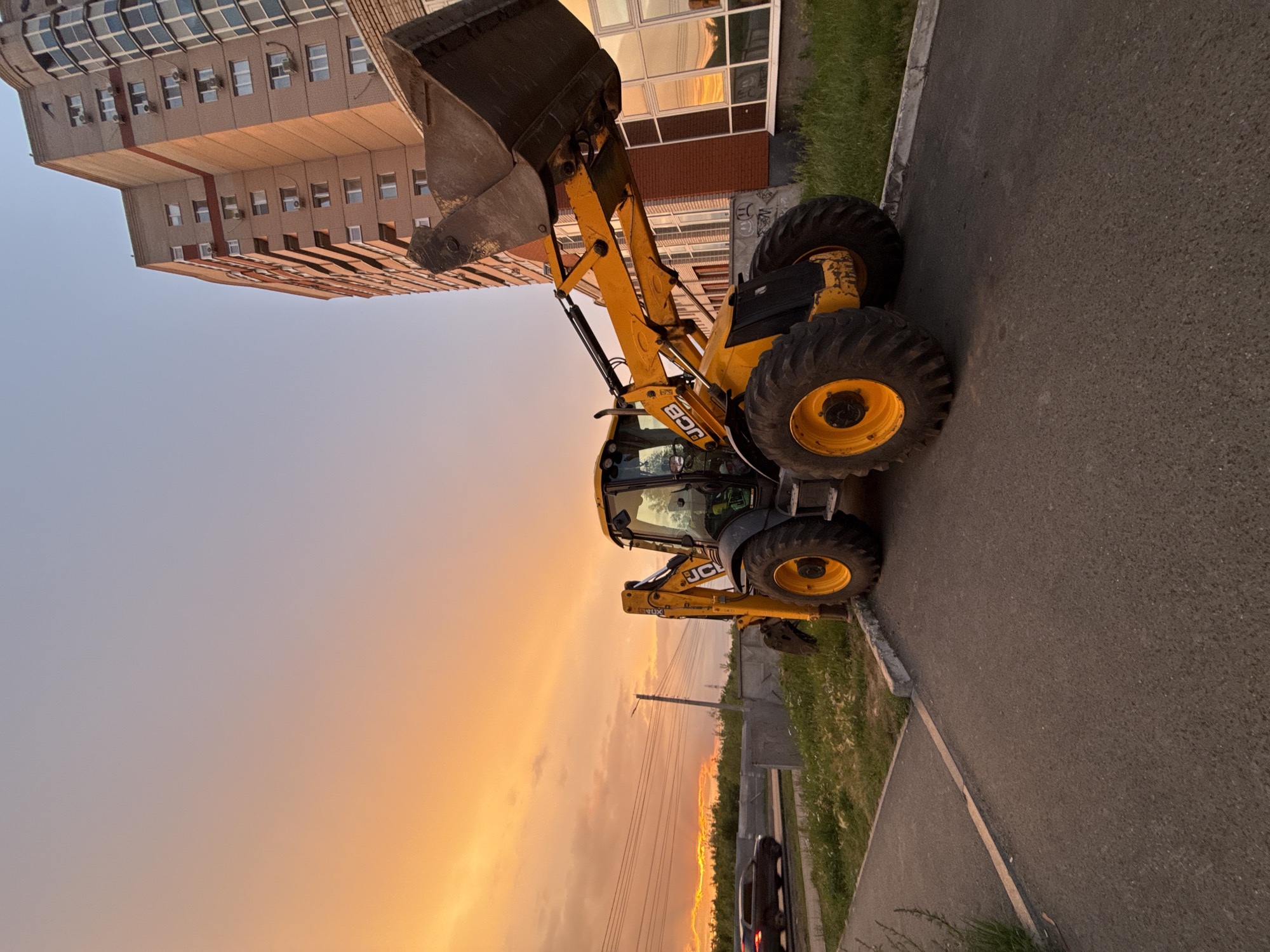 In Izhevsk, Russia, a yellow JCB backhoe loader is parked on a paved area adjacent to a grassy strip and a sidewalk, with a tall, modern brick apartment building on the right. The building features numerous windows and air conditioning units. In the background, a dark-colored car is visible on a road, and a utility pole with overhead wires stands to the left. The sky is filled with dramatic clouds painted in hues of orange, yellow, and pink, indicative of a sunset. The overall scene suggests an urban construction or industrial setting during twilight.