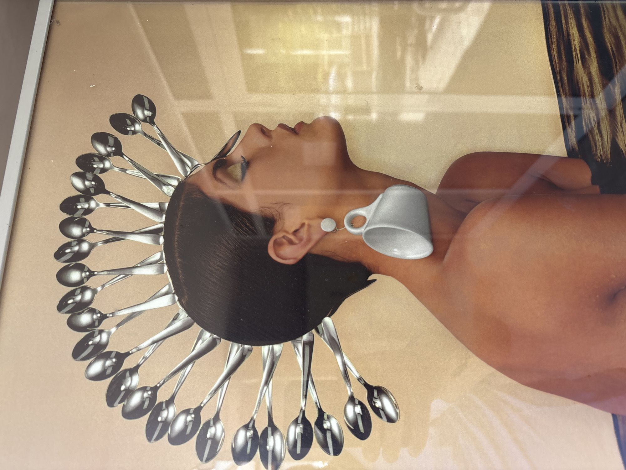 The image is a framed collage featuring a close-up of a woman's profile, adorned with spoons arranged in a semicircular crown-like fashion around her head. She wears a large, dangling earring shaped like a white teacup. Her skin appears bronzed, and her makeup includes dramatic eyeliner and a touch of eyeshadow. The background is a light beige with subtle textured lines and a suggestion of light filtering from a window on the right. The bottom of the collage depicts a dark, textured area resembling water or wood grain. The overall impression is one of surreal and artistic portraiture. The photograph was taken in Plovdiv, Bulgaria.