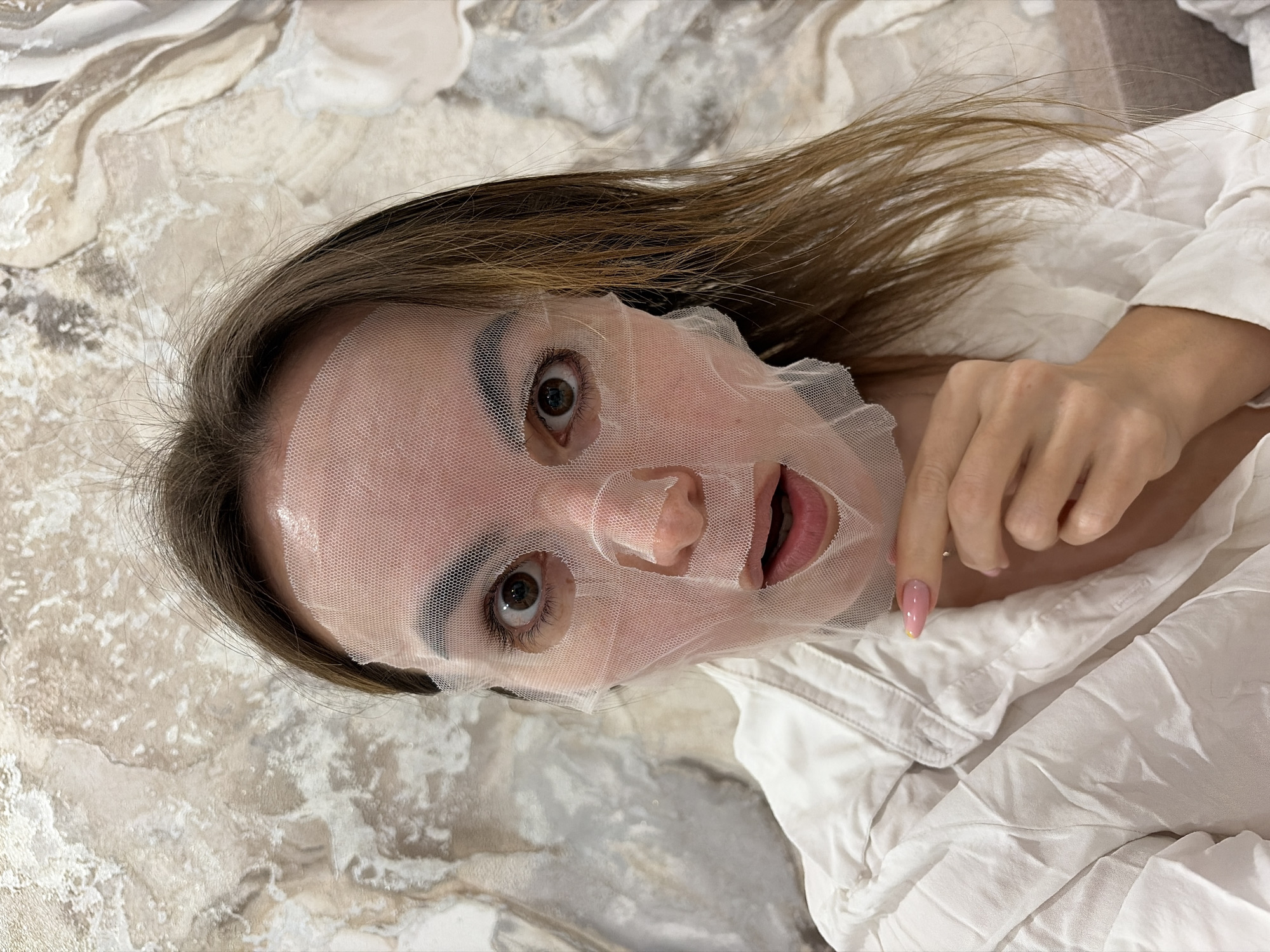 A woman with brown hair and a transparent sheet mask on her face lies on a surface with a marble-like pattern. She is wearing a white shirt. Her hand is visible, with one fingernail showing a pink and yellow design. The setting is indoors. There is no indication that the image was taken in Izhevsk, Russia.