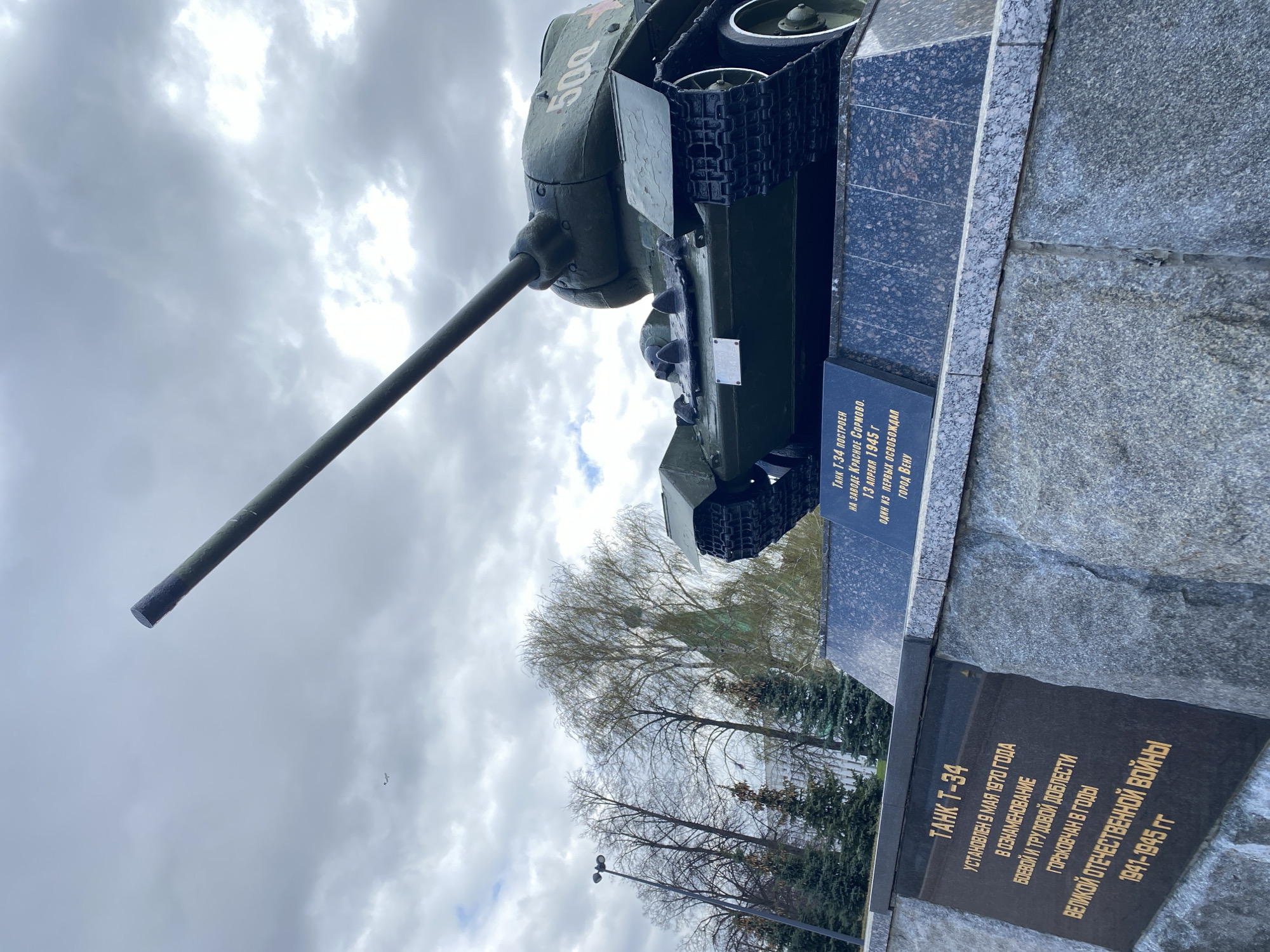 The image displays a monument featuring a Soviet T-34 tank, set outdoors under a cloudy sky. The dark green tank, prominently marked with the number "500" in white on its turret, is mounted on a multi-tiered pedestal made of dark, polished stone. Its long main gun barrel extends towards the upper left of the frame.
Two plaques with gold Cyrillic lettering are affixed to the pedestal. The lower, larger plaque reads: "TANK T-34. INSTALLED MAY 9, 1970, IN COMMEMORATION OF THE COMBAT AND LABOR VALOR OF GORKY CITIZENS DURING THE YEARS OF THE GREAT PATRIOTIC WAR 1941-1945." The upper plaque states: "TANK T-34 WAS BUILT AT THE KRASNOYE SORMOVO FACTORY. ON APRIL 13, 1945, IT WAS ONE OF THE FIRST TO LIBERATE THE CITY OF VIENNA." This context indicates the monument is located in Nizhny Novgorod (formerly Gorky), Russia, celebrating local contributions to World War II.
The sky is heavily overcast with bright clouds, suggesting a daytime scene with diffused lighting. In the background, a variety of trees are visible, some deciduous with bare branches or early foliage, and some evergreen. A street lamp post can be seen on the left side, adding to the urban park setting. The image captures a static historical memorial.
