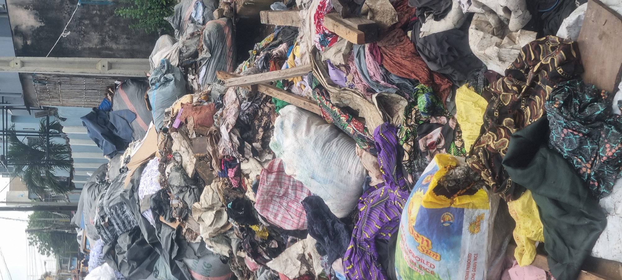 In Ikeja, Nigeria, a massive pile of refuse is located next to a wall, with a street and buildings in the background. The refuse consists of various items, including large bags, textiles of different colors and patterns, and what appears to be construction debris such as broken concrete and wood. Some of the textiles are clothing items, some are in large sacks, and others are spread out. A large bag of "Golden Sun Indian Parboiled Long Grain Rice" is visible in the foreground. No people are clearly visible in the image, though there may be individuals in the distant background on the street. The environment appears to be an urban setting, characterized by the surrounding buildings and the presence of a street.